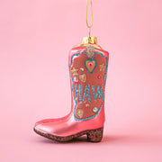 On a pink background is a glass ornament in the shape of pink cowgirl boots with blue pink and gold detailing along with glitter text that reads, "Yee" on one side and "Haw" on the other.