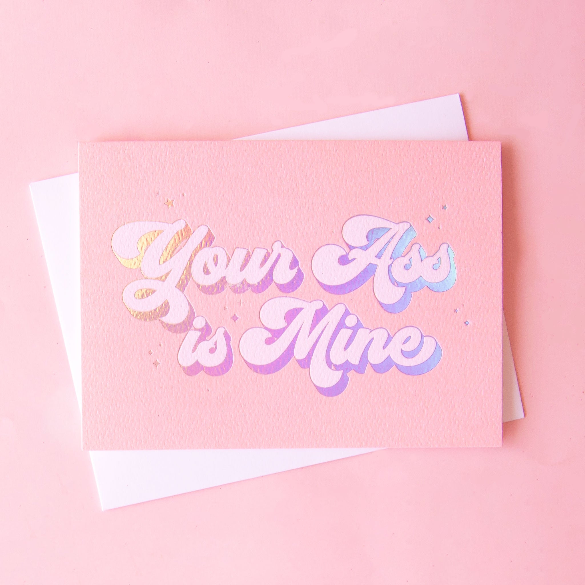 A pink card with a white envelope and holographic text that reads, 'Your Ass is Mine'. 