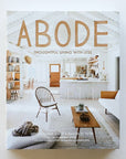A book lays on a table titled "Adobe - thoughtful living with less." Cover shows inside a bright country - boho style home.
