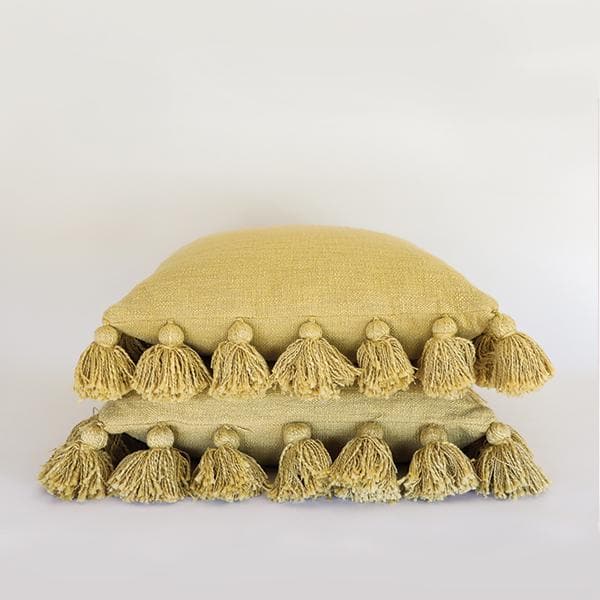 Against a white background is a stack of two yellow, square pillows. The edge of both pillows are lined with small yellow balls with tassels. 