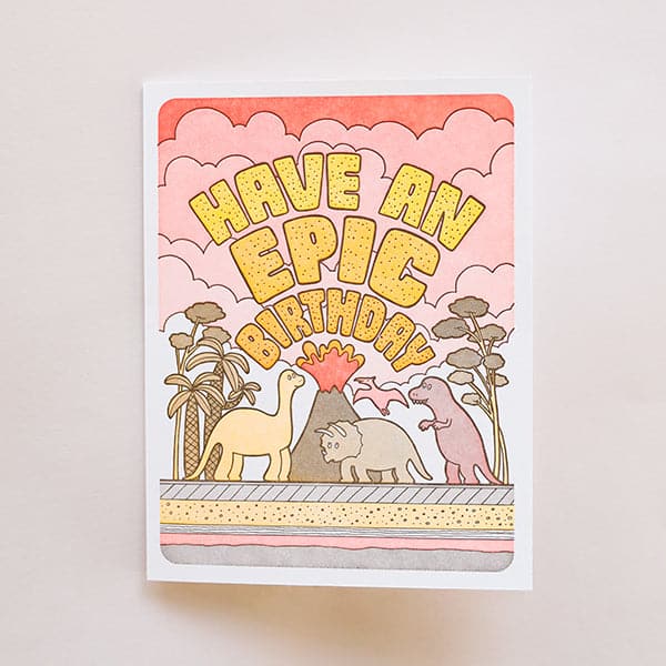 On a white background is a yellow and orange card with dinosaurs and a volcano graphic along with text arched over the illustration that says, "Have An Epic Birthday" along with a coordinating yellow envelope.