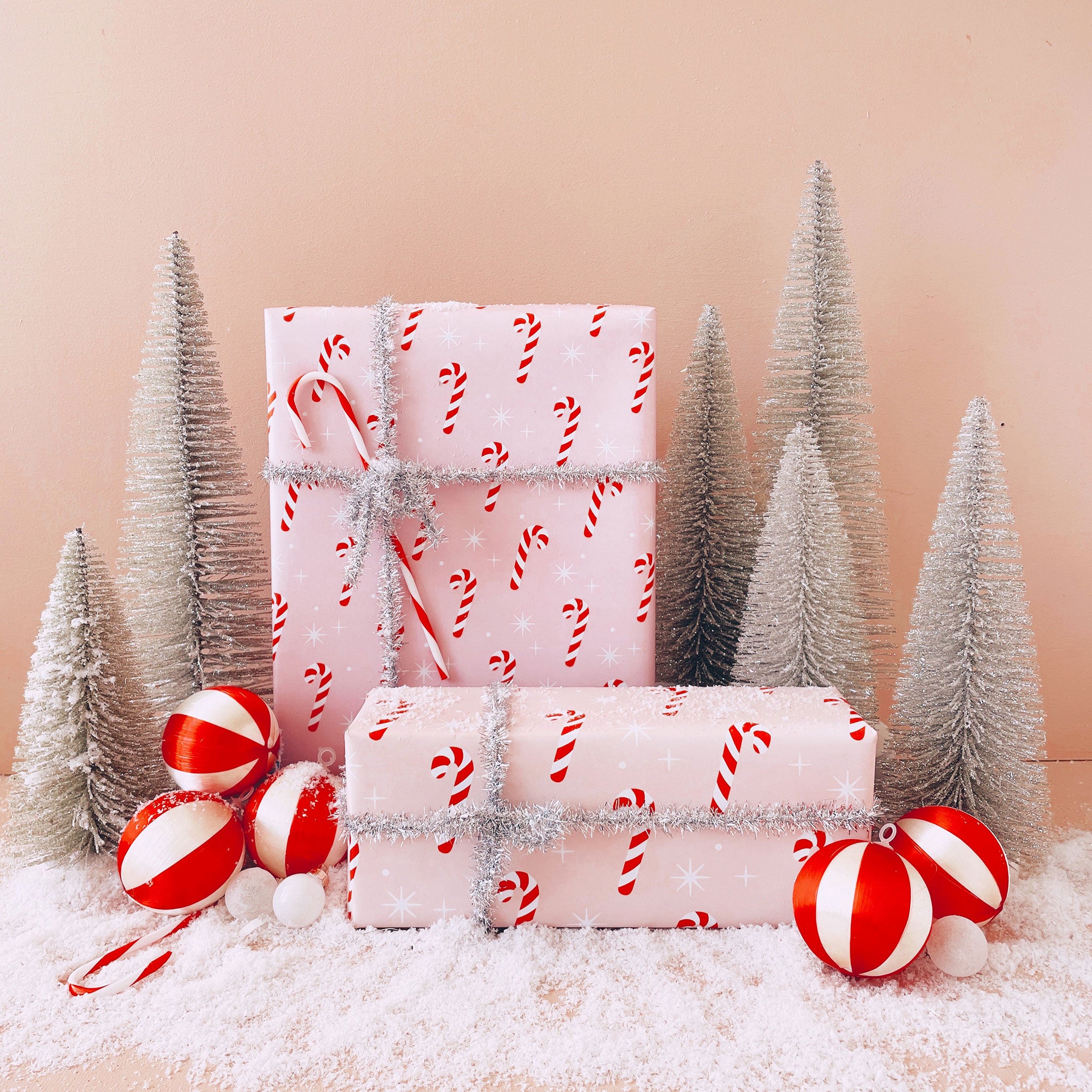 A single sheet of gift wrap with a blush background and a repeating red and white candy cane print. The gift wrap is reversible and the other side features a light purple / lavender background with an all over red and white candy cane design but the candy canes are slightly smaller than the reverse. Present is wrapped with a silver tinsel ribbon and surrounded by snow, red and white ornaments and silver tinsel trees.