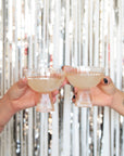 Two light peach beveled coupe glasses with a ribbed stem and a wider ribbed cup being held up doing a cheers in front of a silver tinsel background.