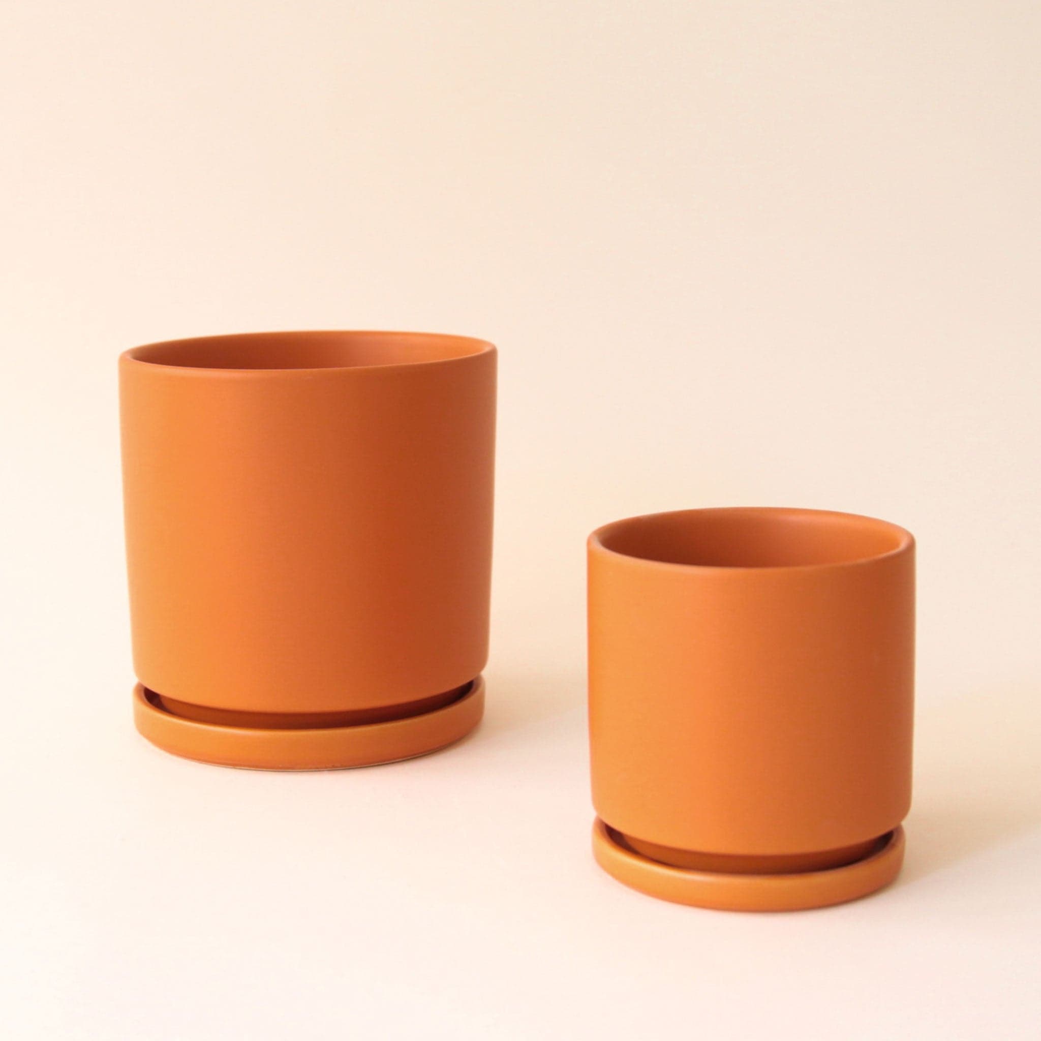 On a cream background is two different sized terracotta ceramic pots with removable trays for watering.