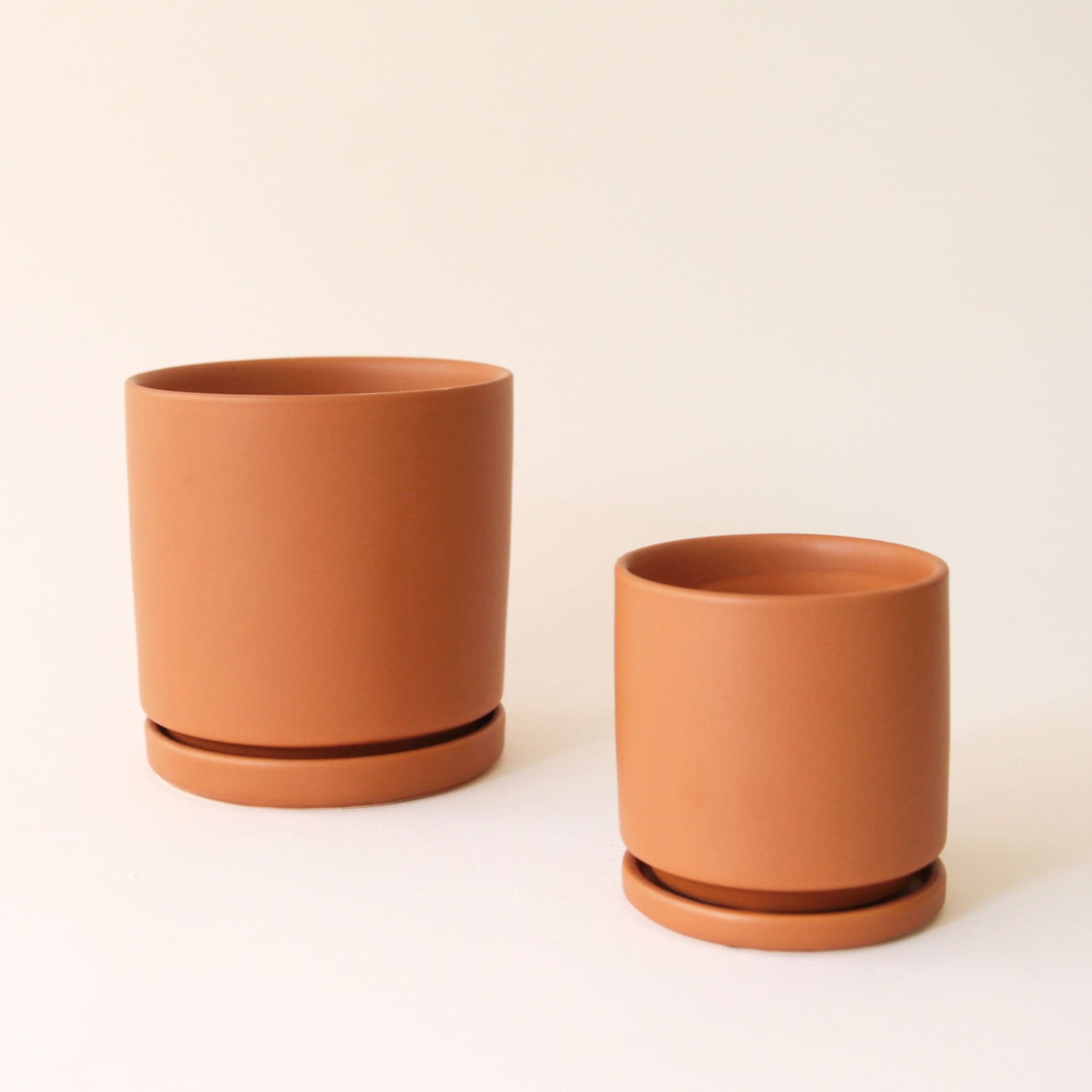 On a cream background is two different sized brownish orange ceramic pots with removable trays for watering.