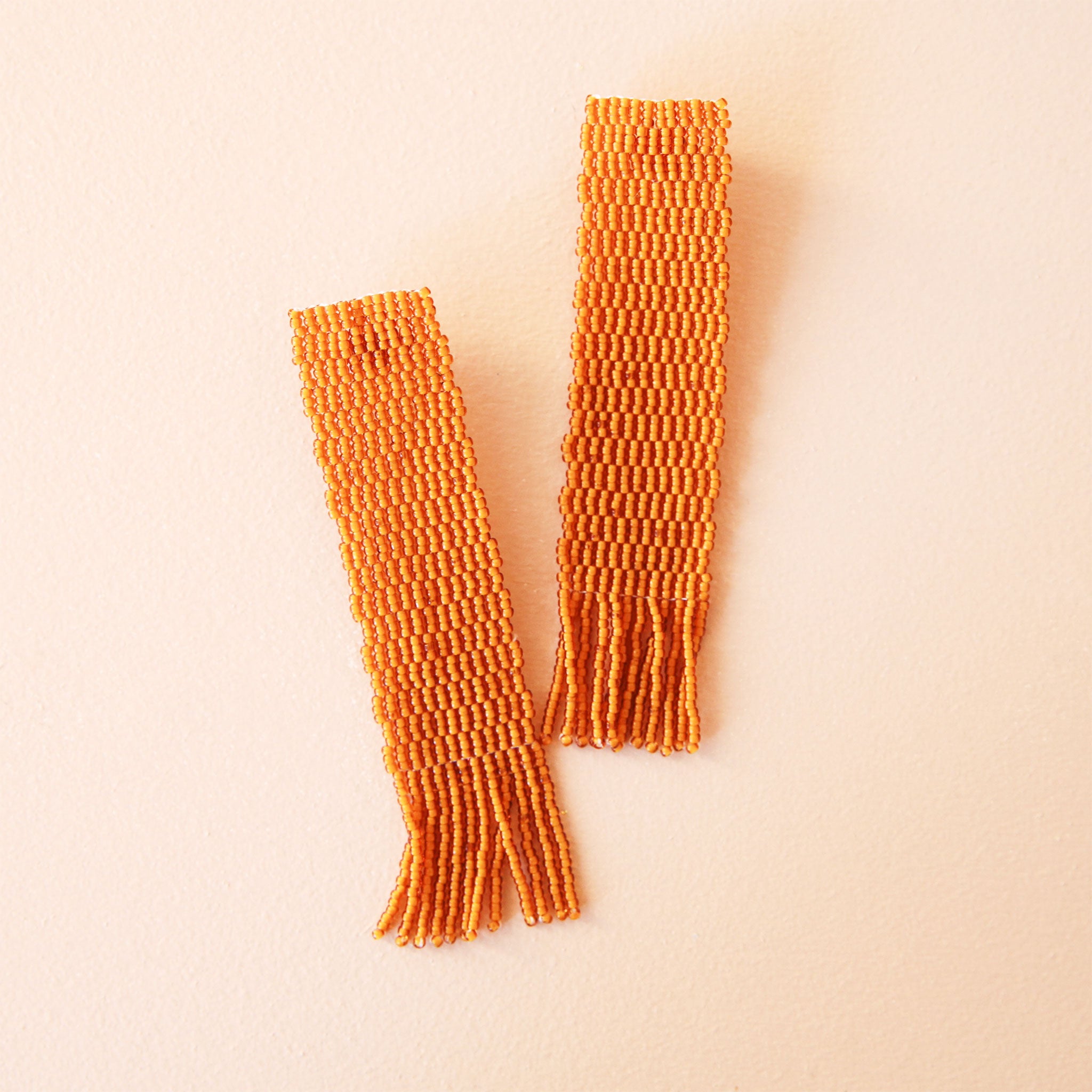 A four inch long by one inch wide strip earring made up of tiny orange, hand beaded beads with a fringe detail at the bottom.