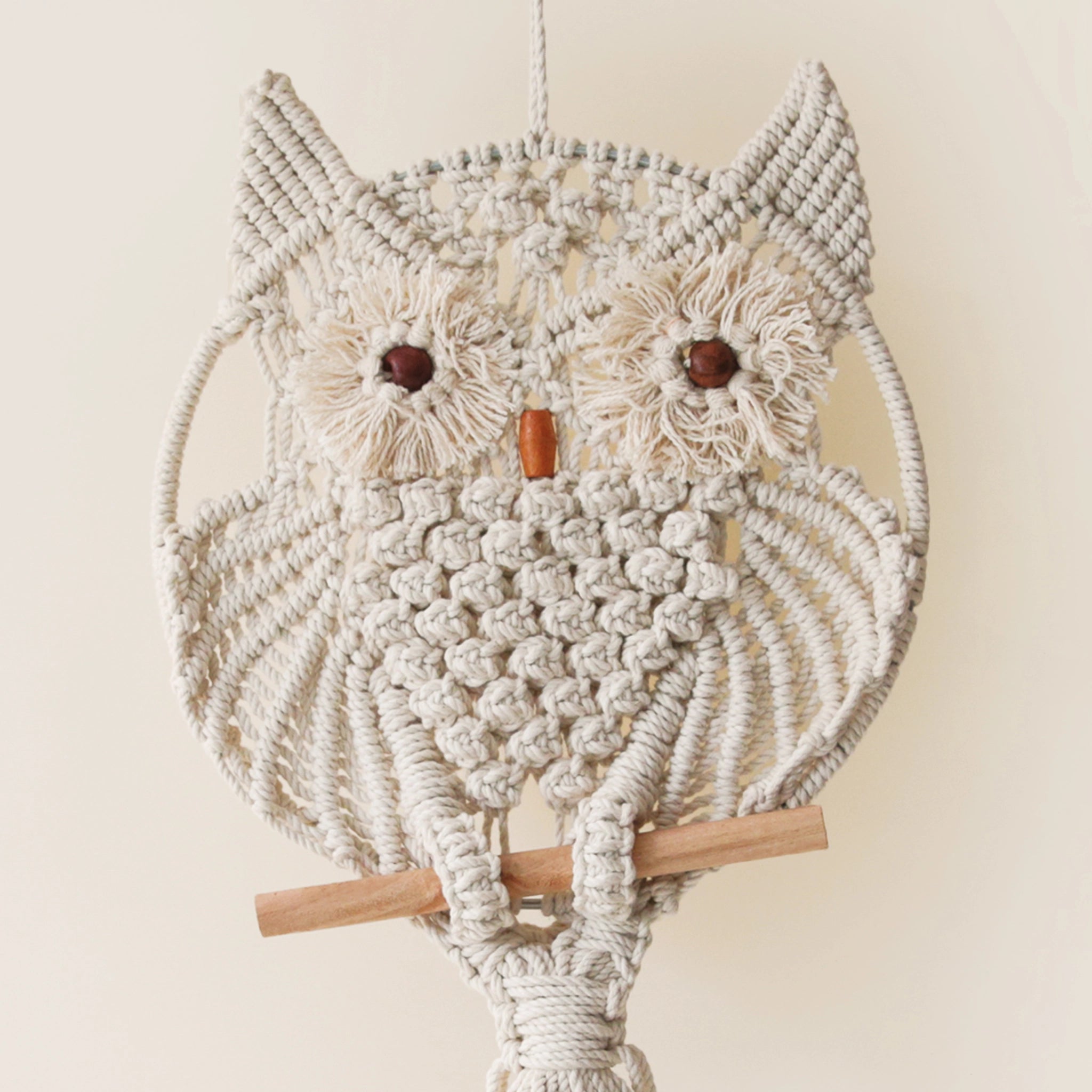 A beige round macrame in the shape of a circle with an owl design along with two brown beads placed for eyes and a terracotta colored bead meant to be its beak.