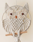 A beige round macrame in the shape of a circle with an owl design along with two brown beads placed for eyes and a terracotta colored bead meant to be its beak.