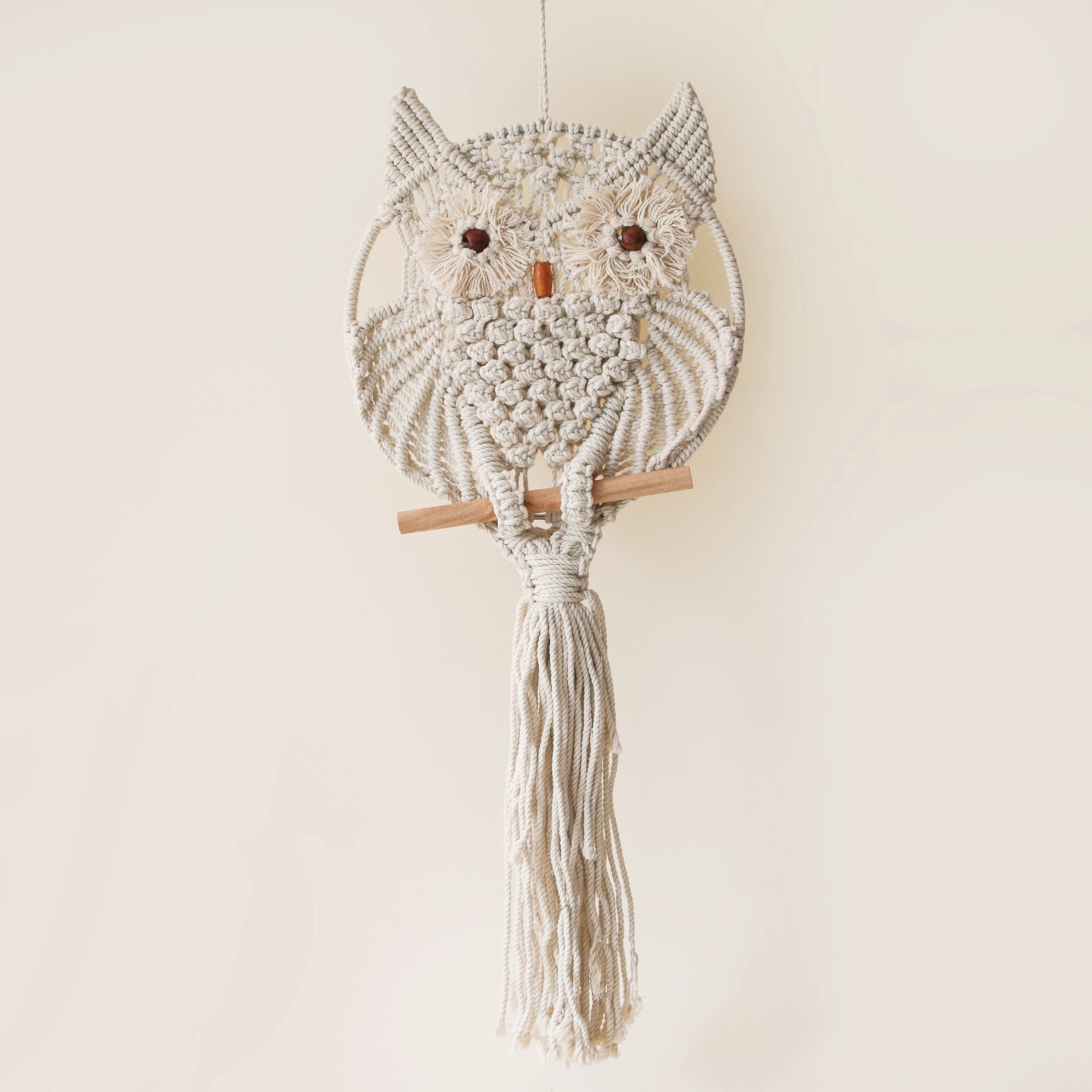 A beige round macrame in the shape of a circle with an owl design along with two brown beads placed for eyes and a terracotta colored bead meant to be its beak. 