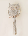 A beige round macrame in the shape of a circle with an owl design along with two brown beads placed for eyes and a terracotta colored bead meant to be its beak. 