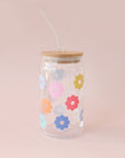 A glass tumbler with multi colored daisies and a wooden lid and a glass straw.