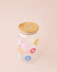 A glass tumbler with multi colored daisies and a wooden lid and a glass straw.