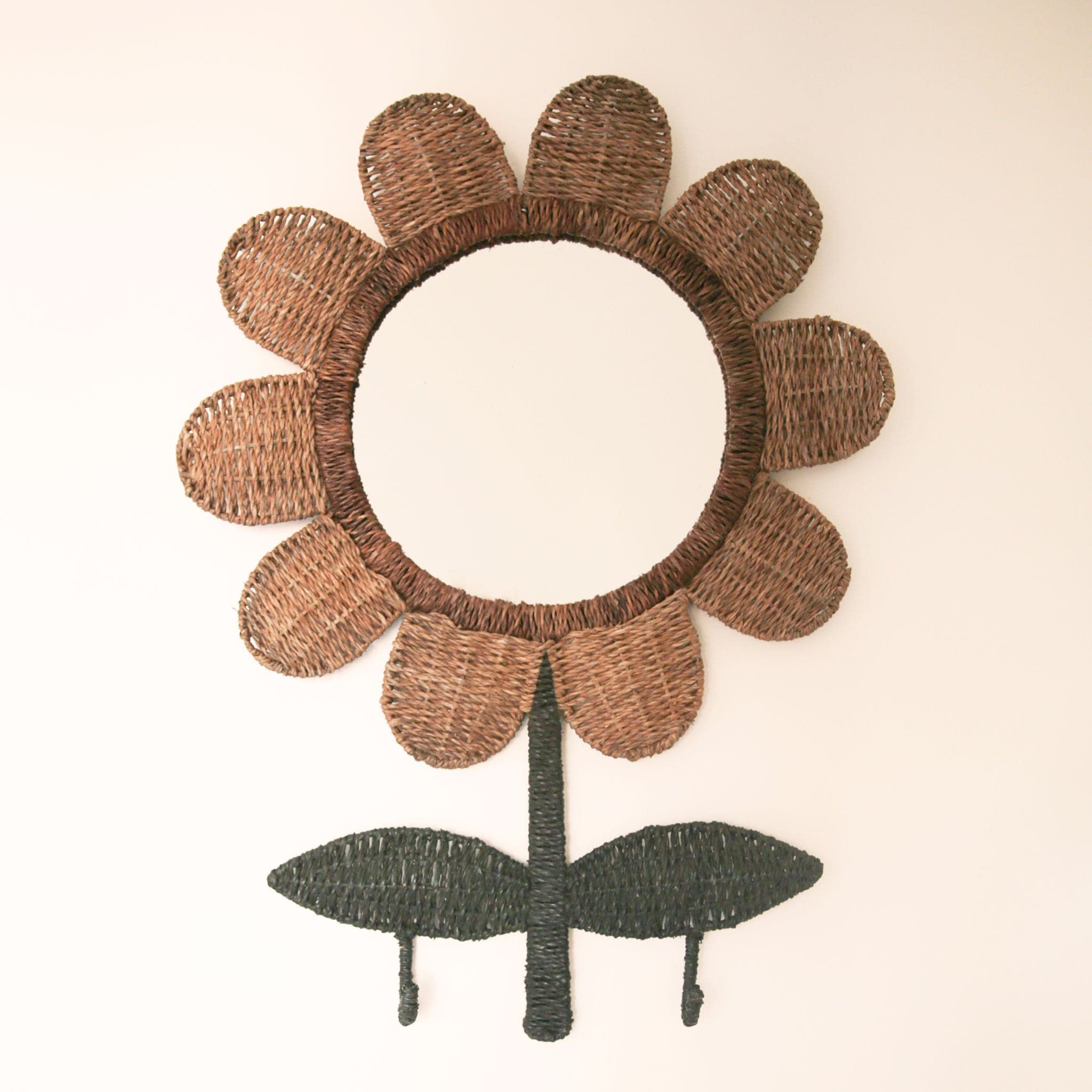 A circle mirror surrounded by woven rattan that is shaped like a flower. The rattan petals are wrapped around the mirror and the 'stem and leaves' are woven green rattan with two small hooks that drop down below the two leaves.