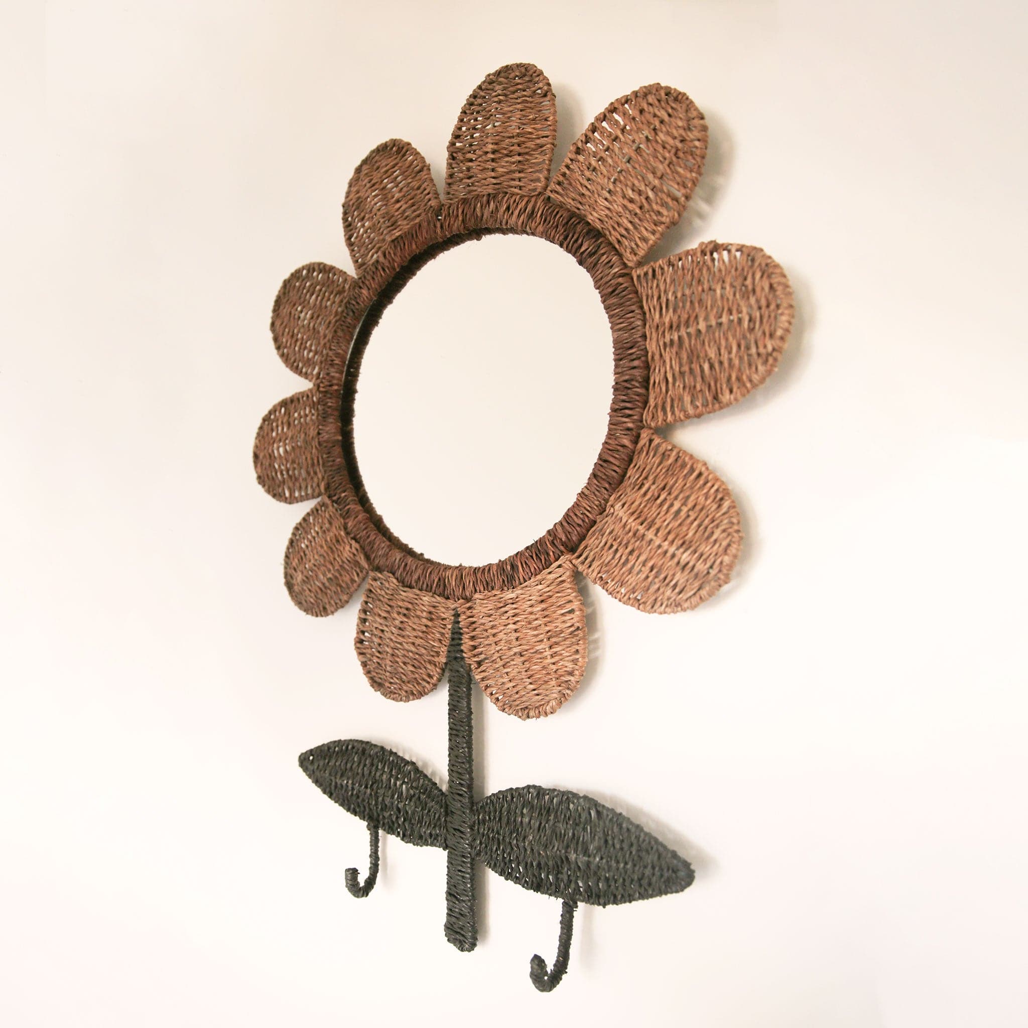 A circle mirror surrounded by woven rattan that is shaped like a flower. The rattan petals are wrapped around the mirror and the 'stem and leaves' are woven green rattan with two small hooks that drop down below the two leaves.