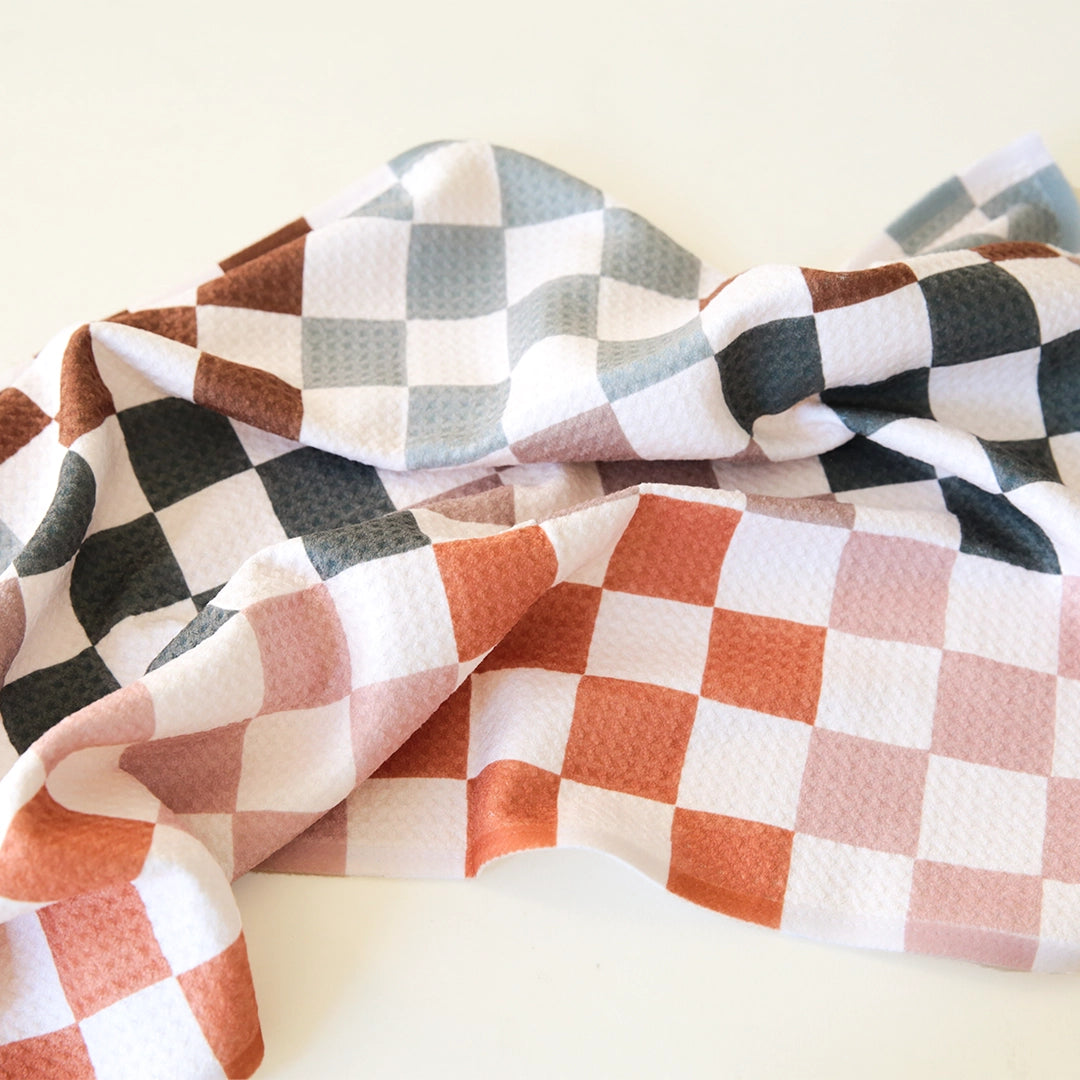 A checkered kitchen towel with different colors of checkers including black, brown, blue/grey, orange and pink.