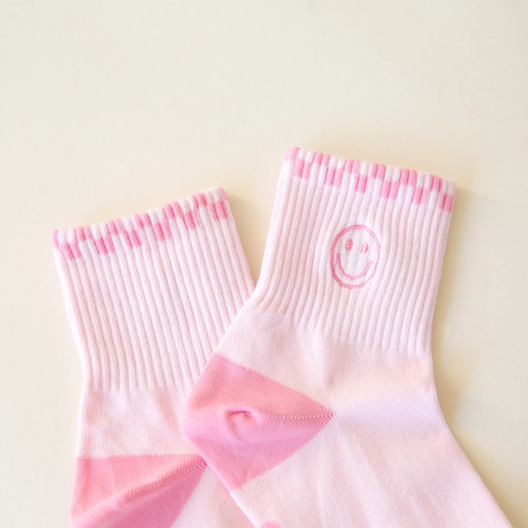 Pink ankle socks that feature a slightly darker pink checkered border across the top of the socks and a smiley face graphic on the ankle.