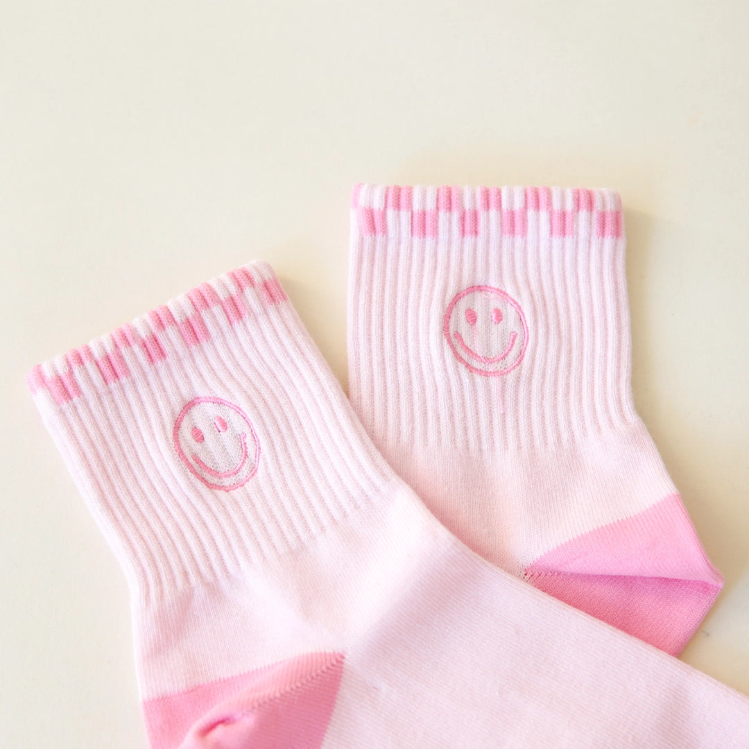 Pink ankle socks that feature a slightly darker pink checkered border across the top of the socks and a smiley face graphic on the ankle.