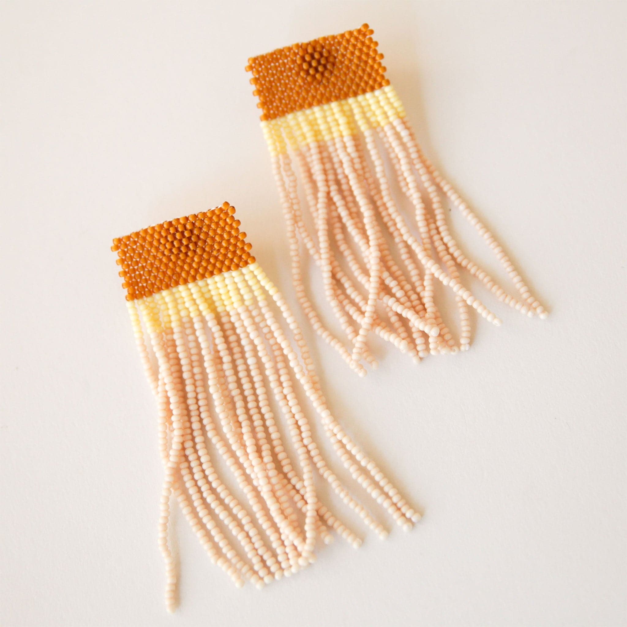 A pair of two beaded earrings with an orange top, a yellow middle and cream beaded strands that dangle.