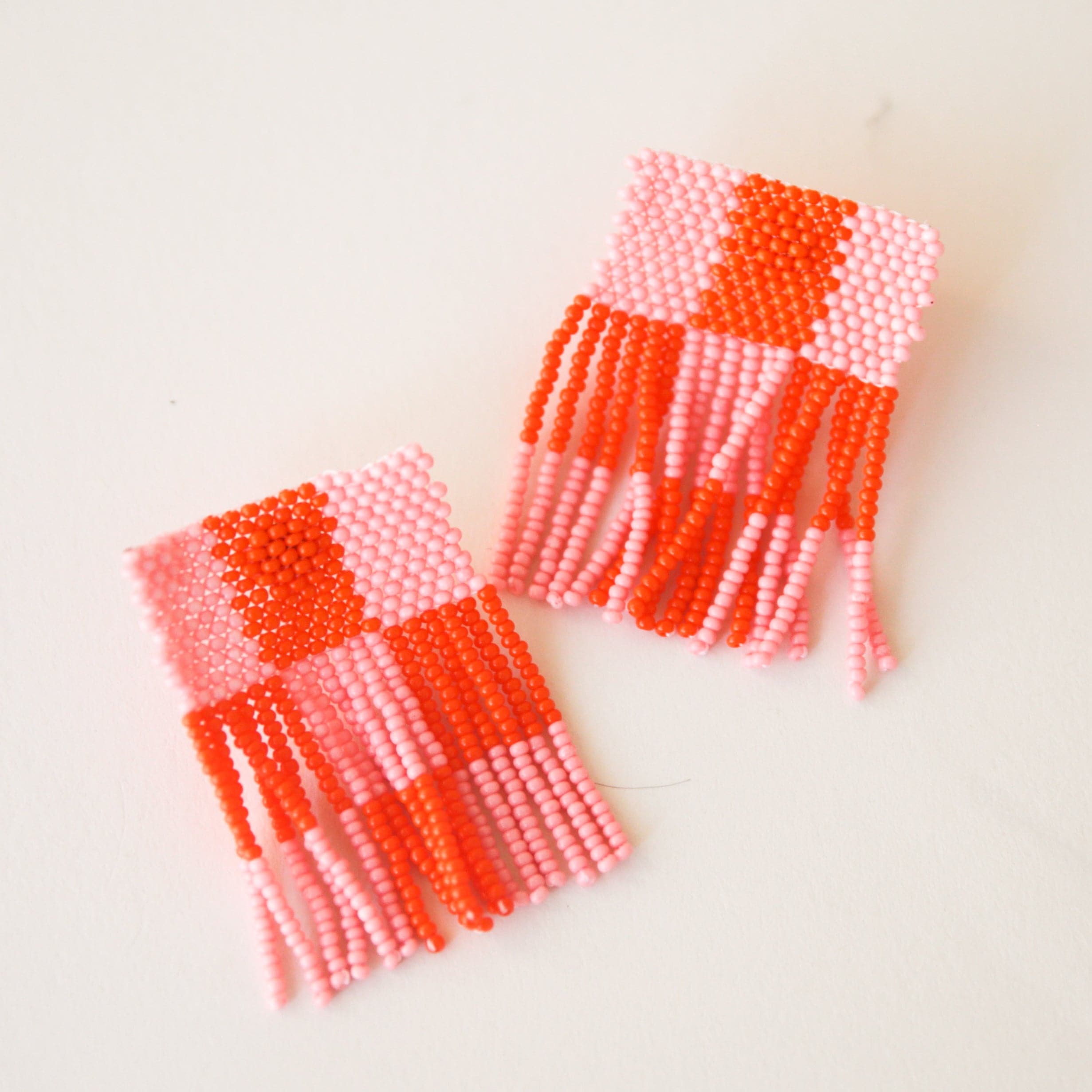 On a cream background is a pair of beaded pink and red checkered earrings with a square shape and a fringed bottom edge. 