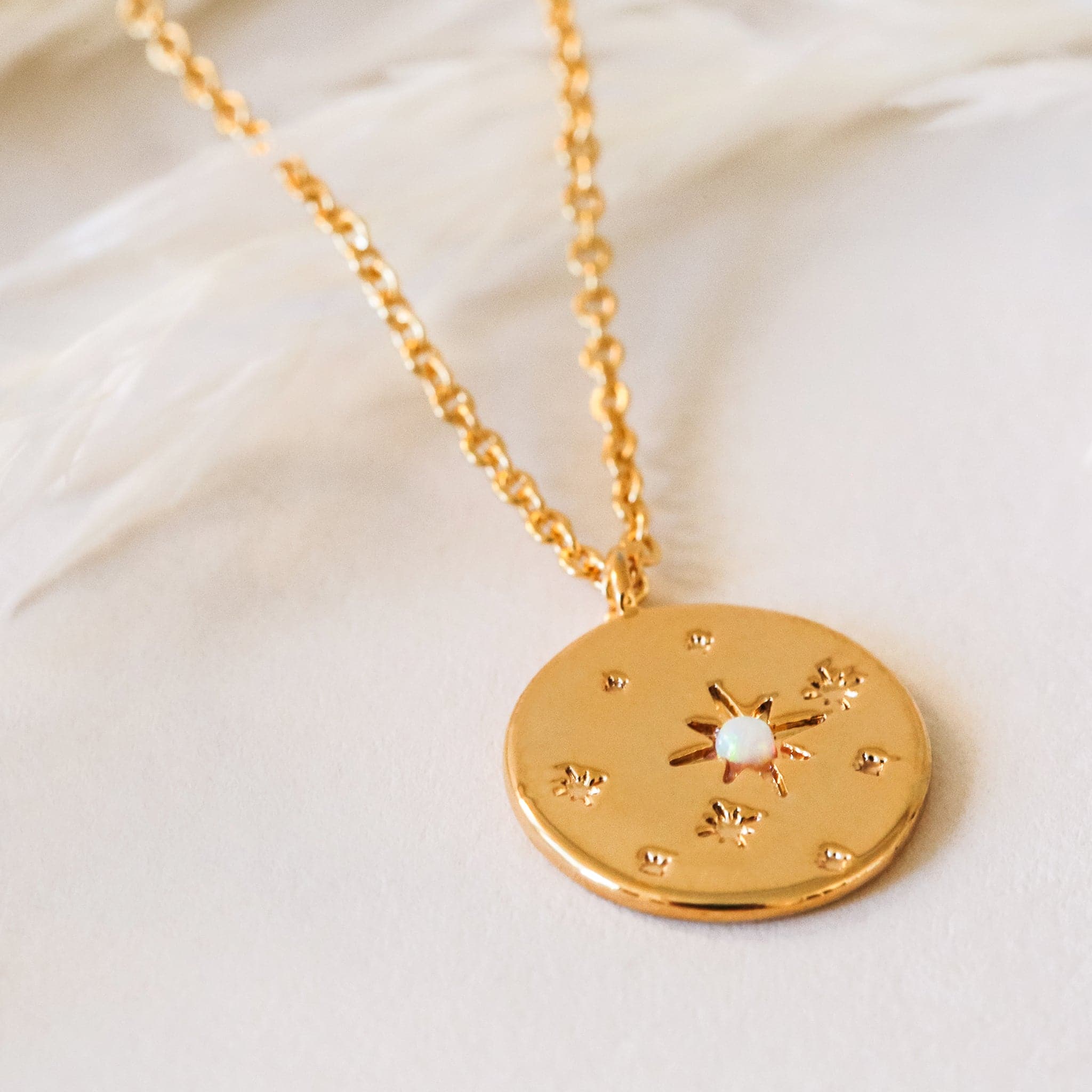 Round star opal necklace in gold. This necklace has a delicate gold chain, with a coin pendant. The pendant has a twinkle star impression with a set opal in the center. This star shape is off-center with other star impressions in a natural display with different sizes scattered across the pendant.