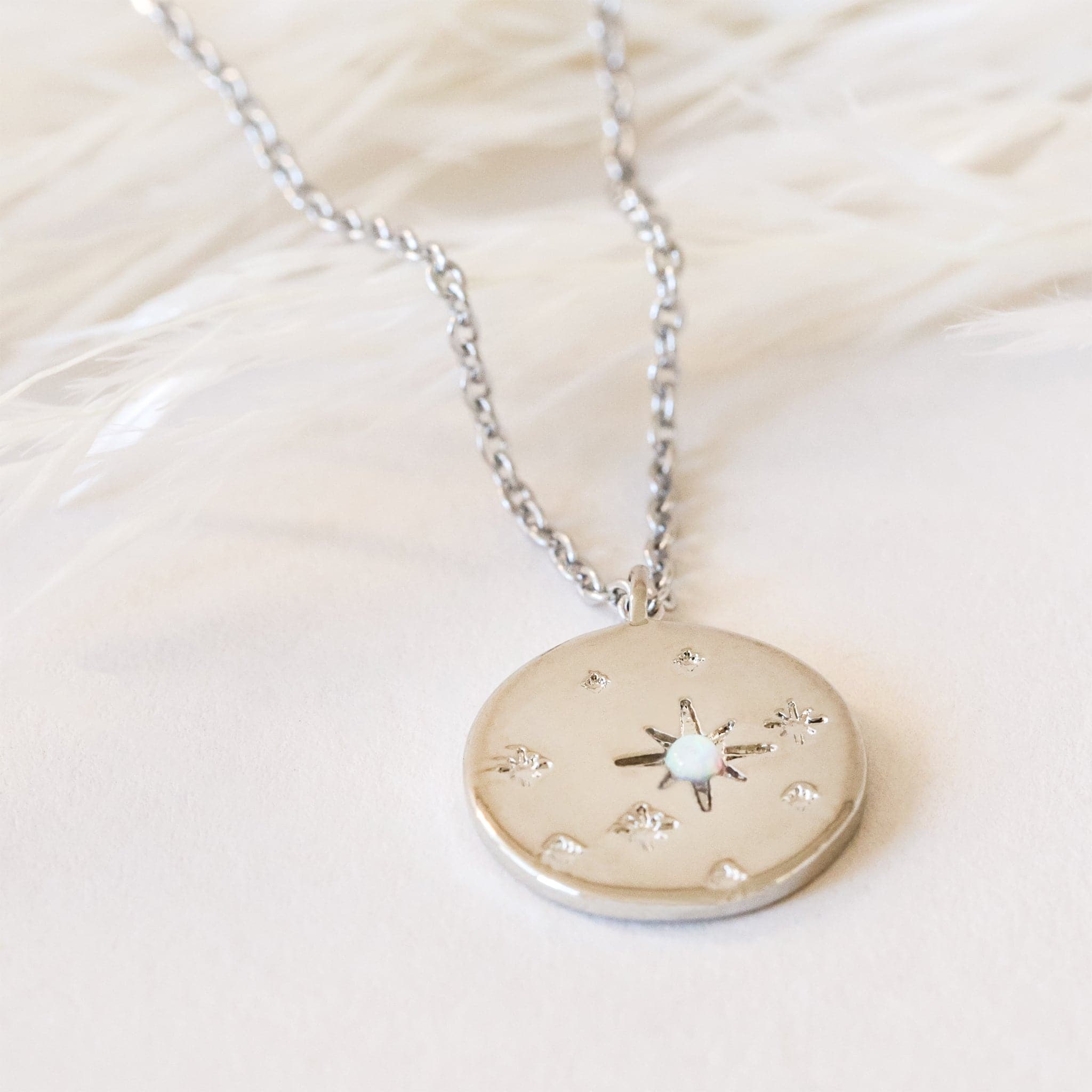 Round star opal necklace in Silver. This necklace has a delicate gold chain, with a coin pendant. The pendant has a twinkle star impression with a set opal in the center. This star shape is off-center with other star impressions in a natural display with different sizes scattered across the pendant.