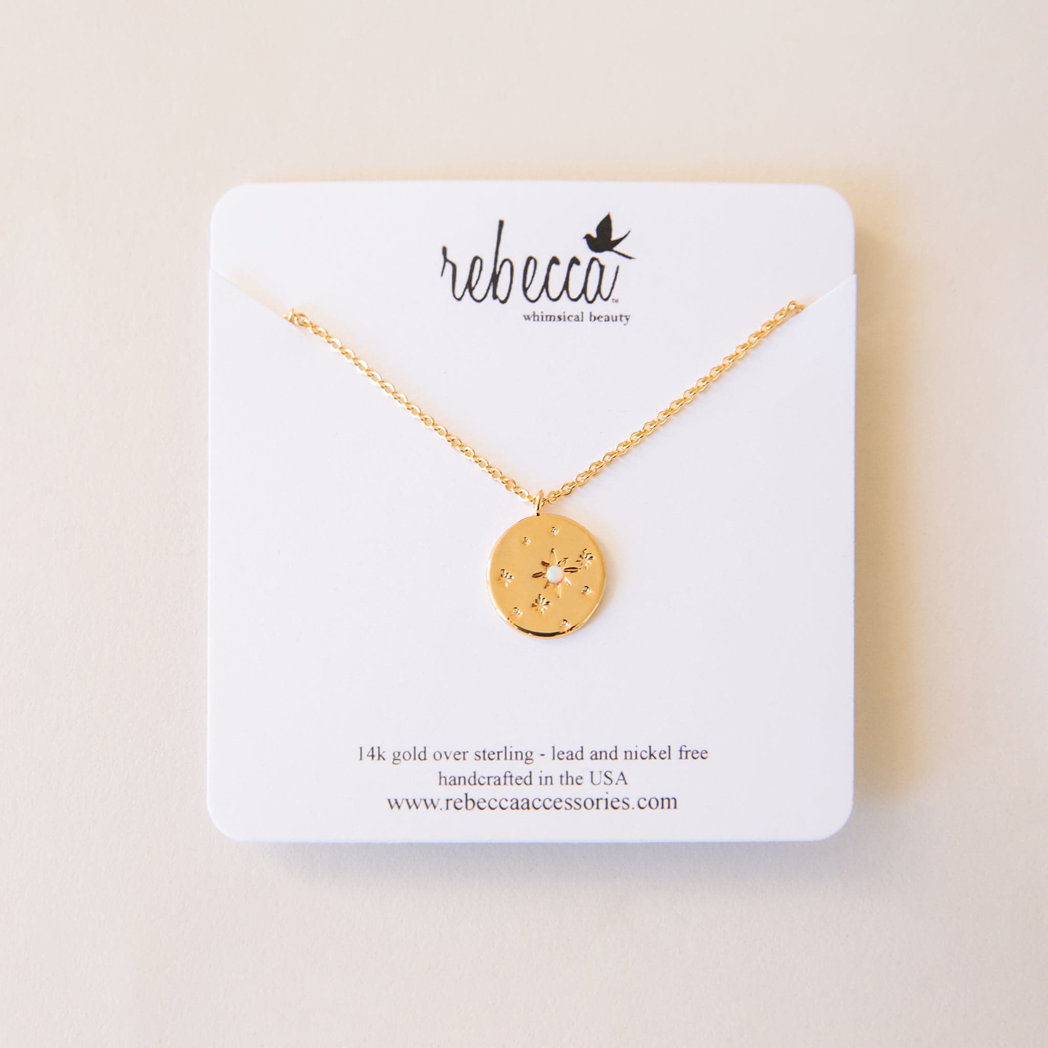 The round star opal necklace on a white card for beautiful gifting and display. The Round star opal necklace in gold. This necklace has a delicate gold chain, with a coin pendant. The pendant has a twinkle star impression with a set opal in the center. This star shape is off-center with other star impressions in a natural display with different sizes scattered across the pendant. 