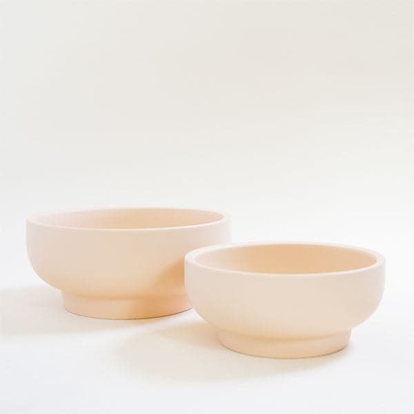 A pair of two pale ivory pedestal bowls, both with tapered bases. To the left is the larger of the pair and to the right sits a slightly shorter bowl. They both are positioned empty besides each other. 