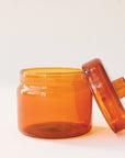 A round glass amber jar with a coordinating glass lid that has a round, flat knob on top.