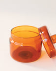 A round glass amber jar with a coordinating glass lid that has a round, flat knob on top.