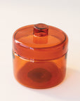 A round glass amber jar with a coordinating glass lid that has a round, flat knob on top.