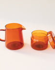 A round glass amber jar with a coordinating glass lid that has a round, flat knob on top photographed next to a matching amber pitcher.