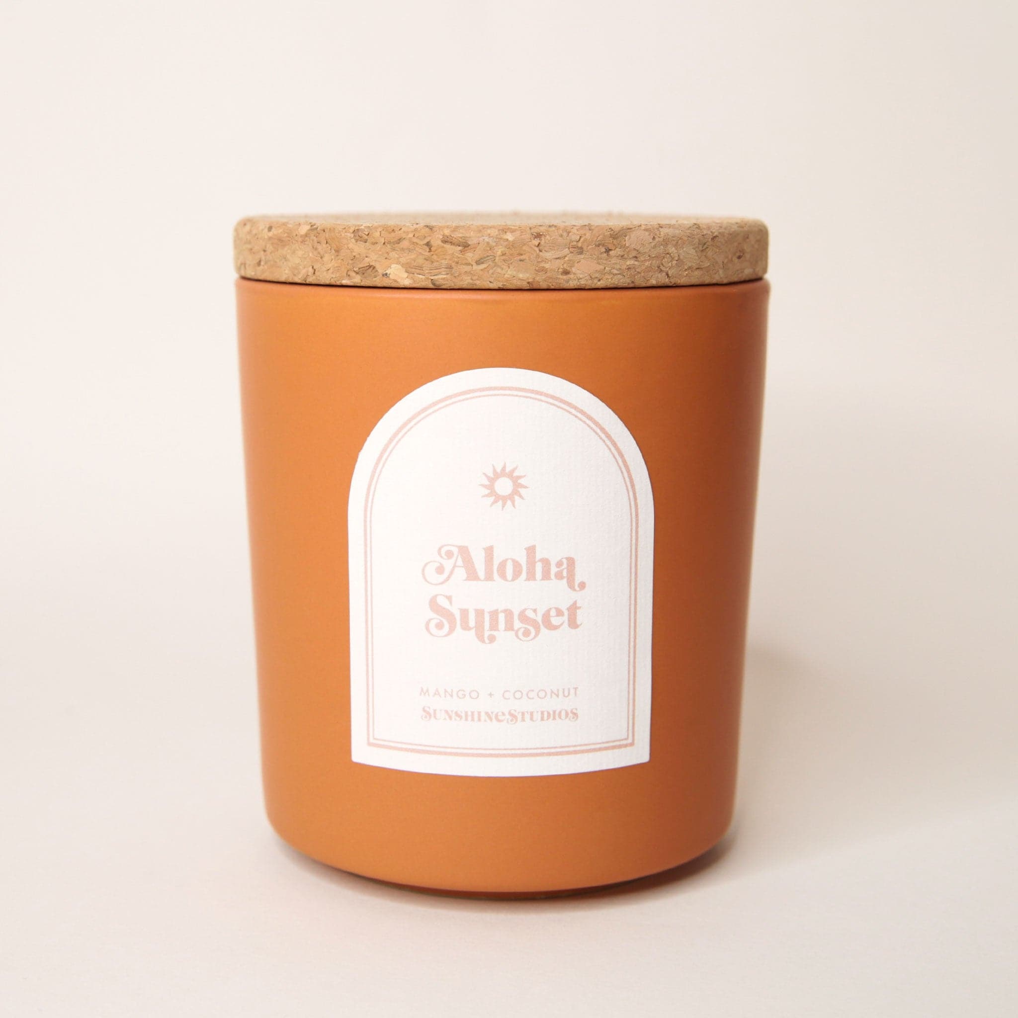 Terracotta orange candle with lid made of cork. An arched label reads 'aloha sunset, mango + coconut, sunshine studios' in soft pink lettering. A small sun design sits above the text. 