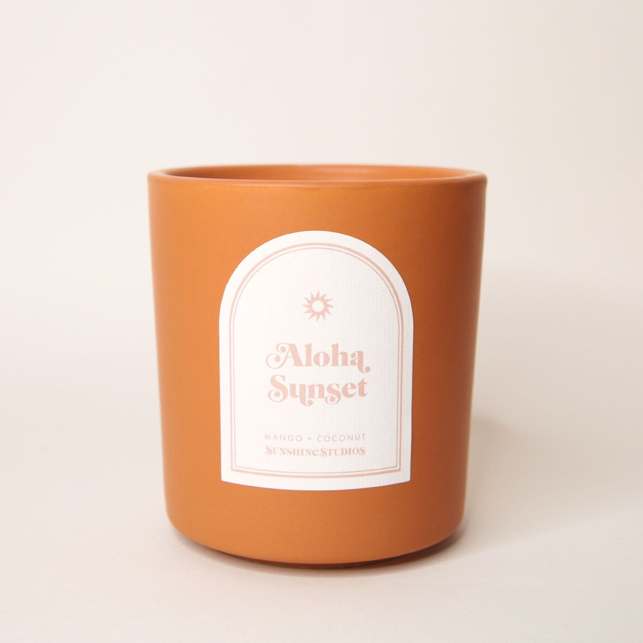 Terracotta orange candle with an arched label that reads 'aloha sunset, mango + coconut, sunshine studios' in soft pink lettering. A small sun design sits above the text. 