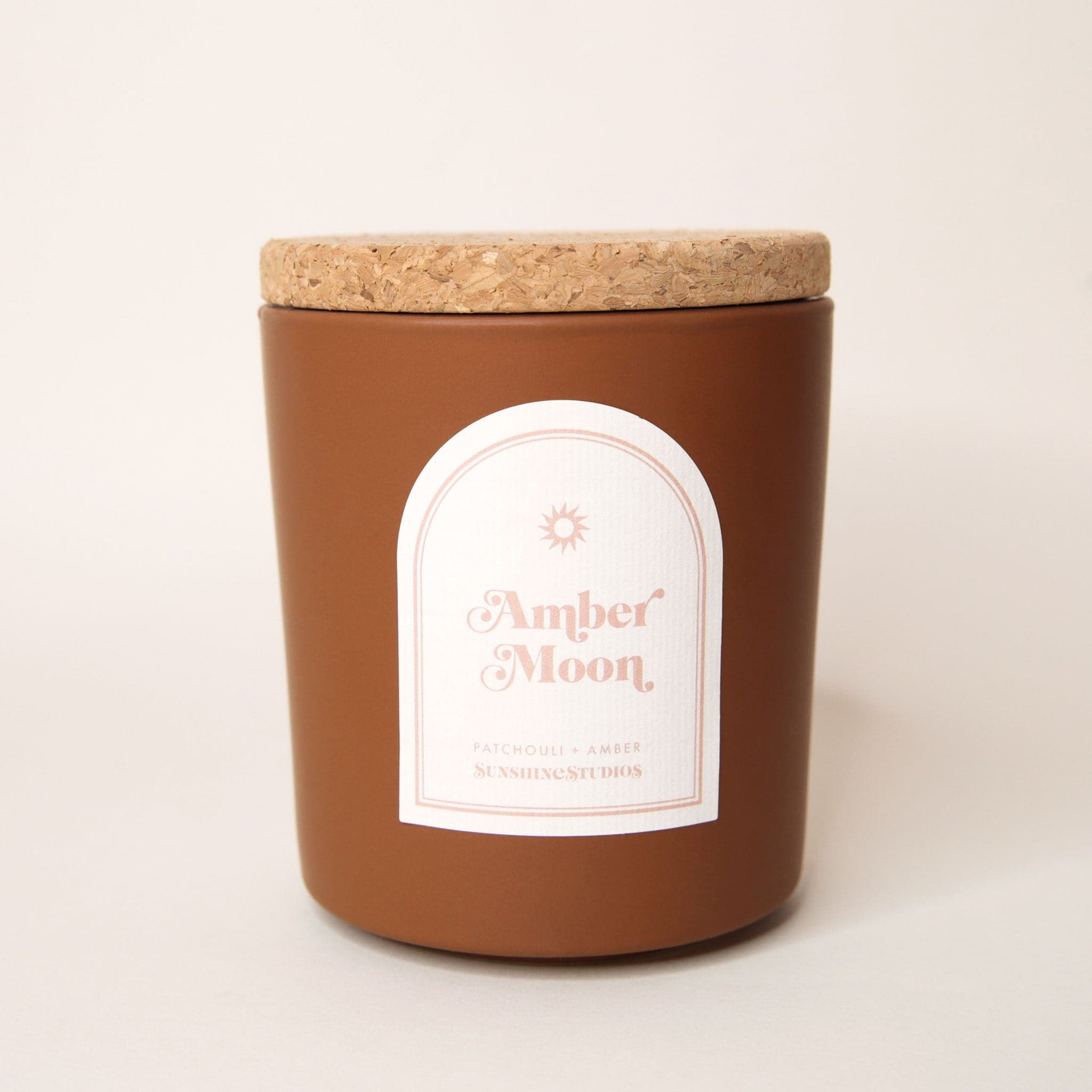 Chocolate brown candle with lid made of cork. An arched label reads 'amber moon, patchouli + amber, sunshine studios' in soft pink lettering. A small sun design sits above the text.