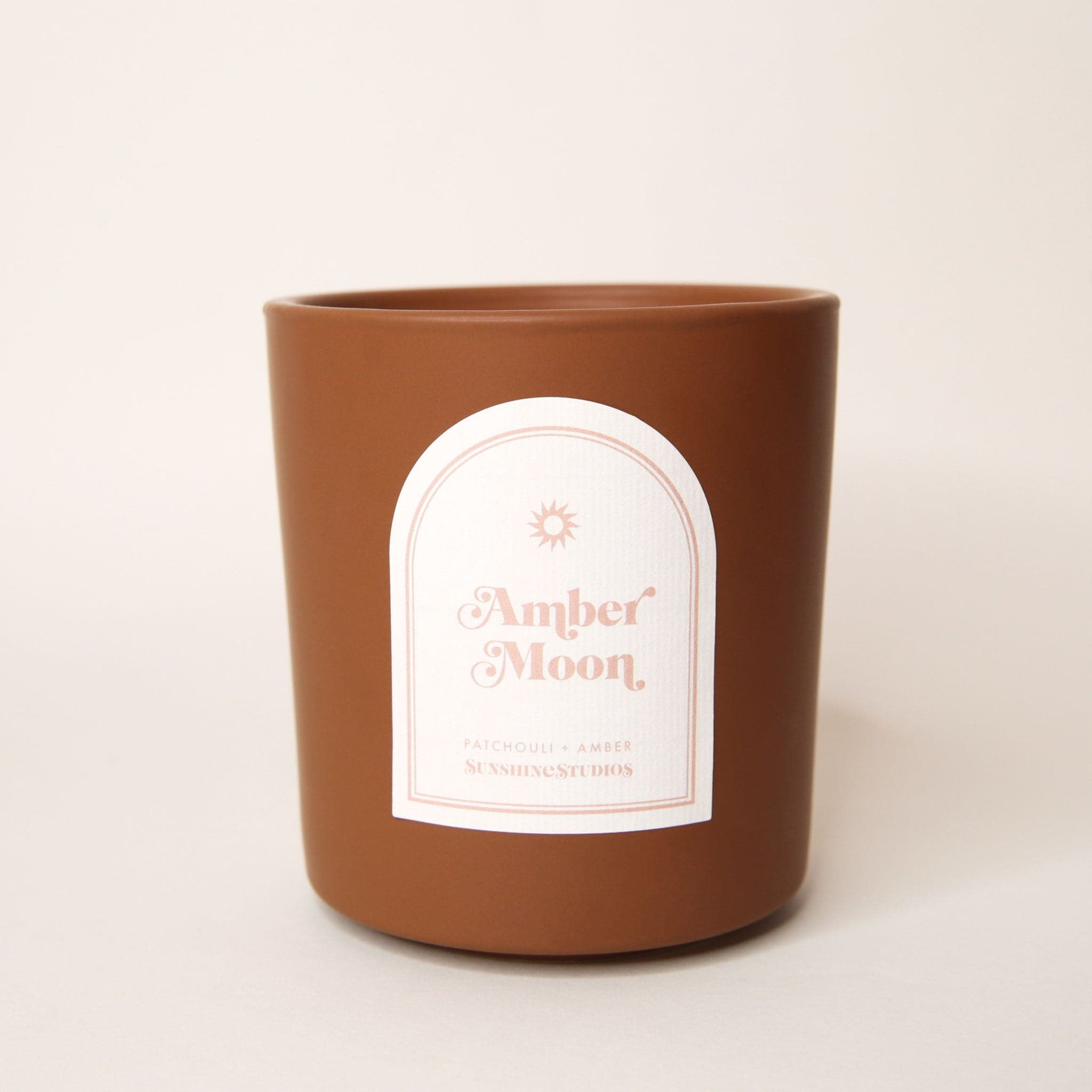 Chocolate brown candle with an arched label that reads 'aloha sunset, mango + coconut, sunshine studios' in soft pink lettering. A small sun design sits above the text.