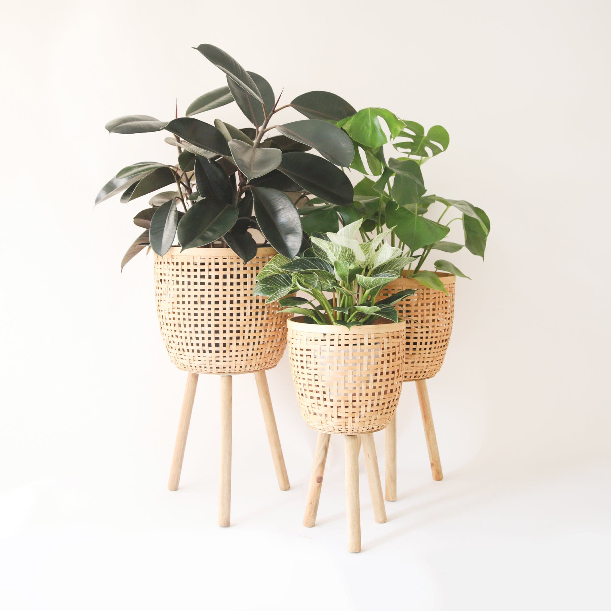 Three different sized, light colored rattan basket plant holders, each standing on three wooden legs. Each basket sold separately.