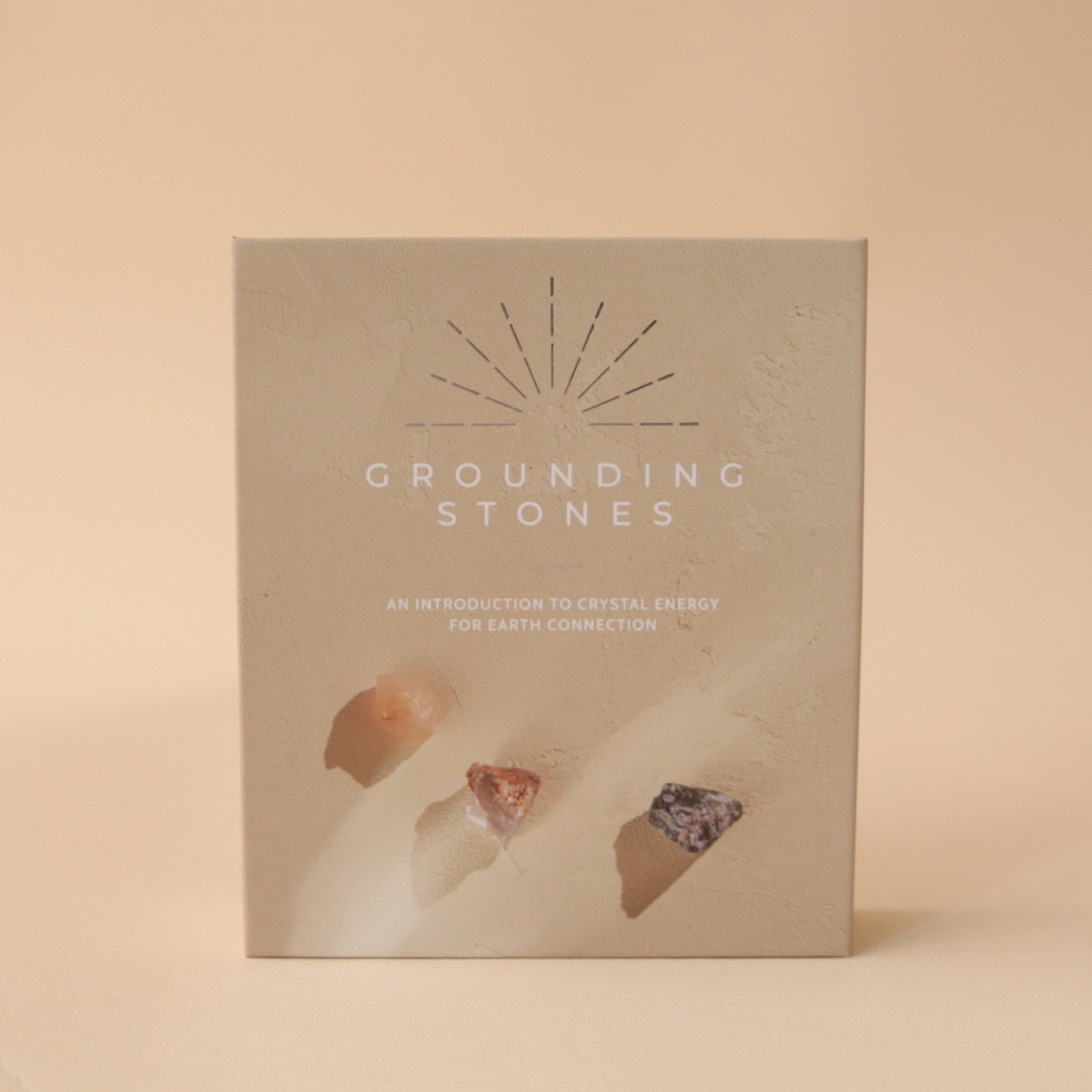 On a light tan background is a tan box that opens up like a book. On the interior is a place for 7 different stones. On the left side of the opening is a list of all the stones and their purpose. The front of the box reads, &quot;Grounding Stones&quot;, &quot;An Introduction To Crystal Energy For Earth Connection&quot;. 