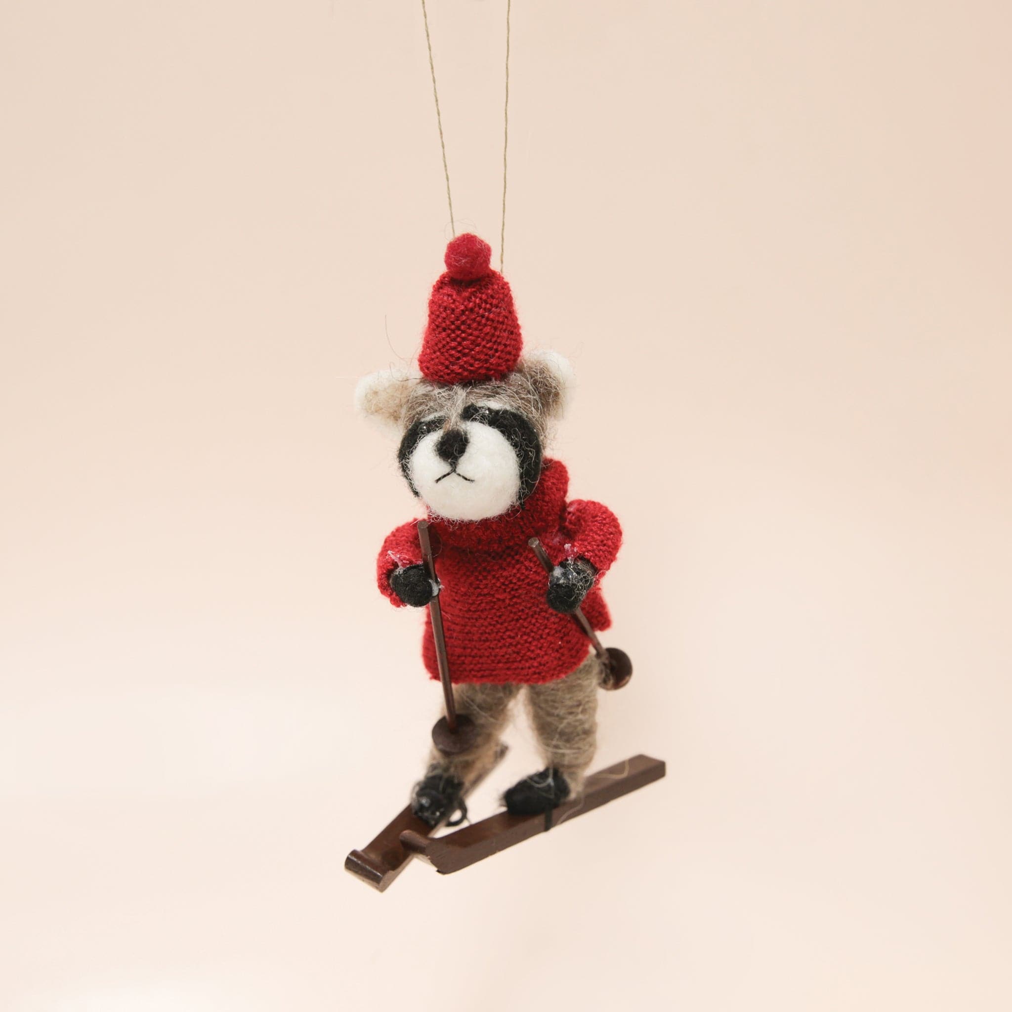 A felt raccoon ornament in a red sweater and hat on brown skis and holding ski poles.