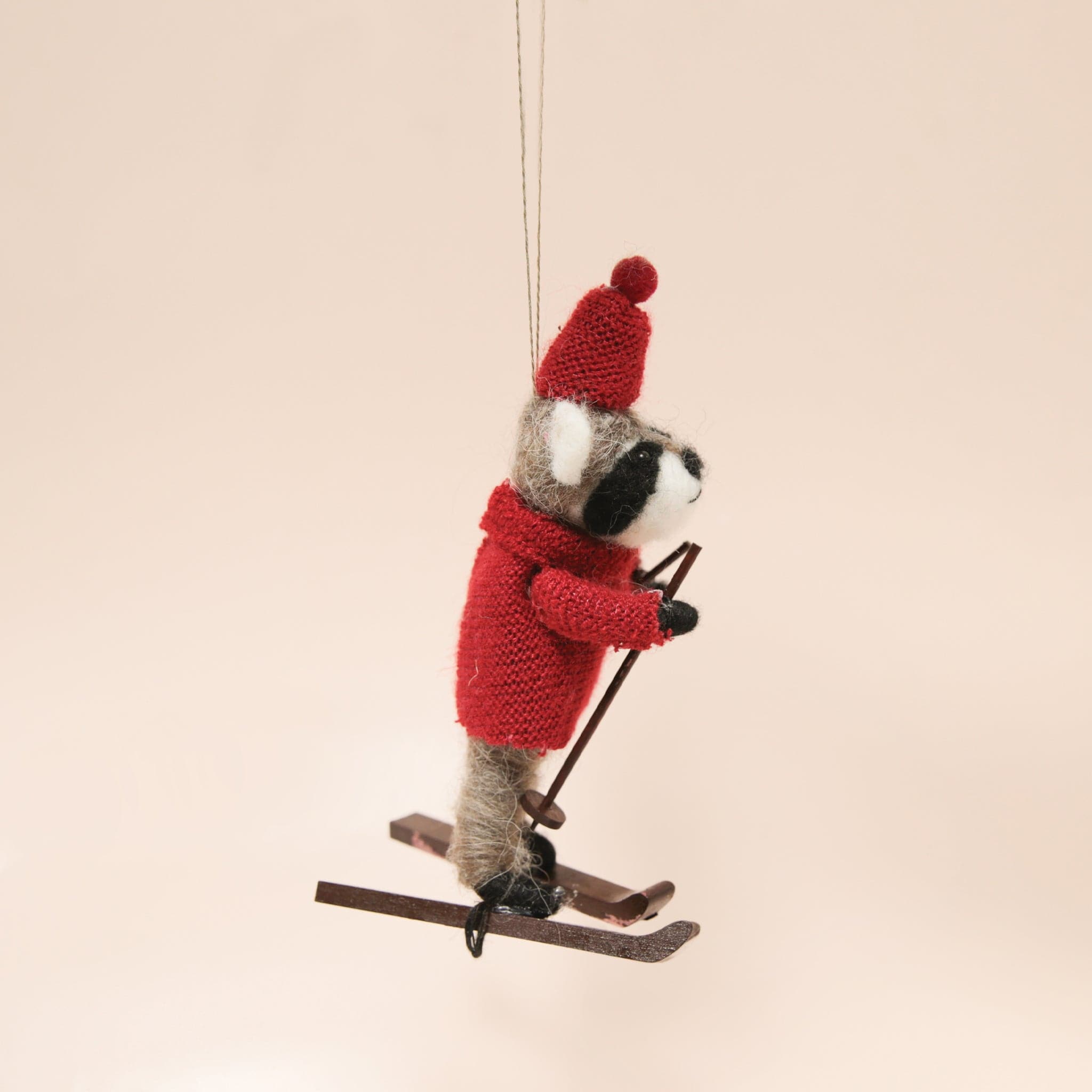 A felt raccoon ornament in a red sweater and hat on brown skis and holding ski poles.