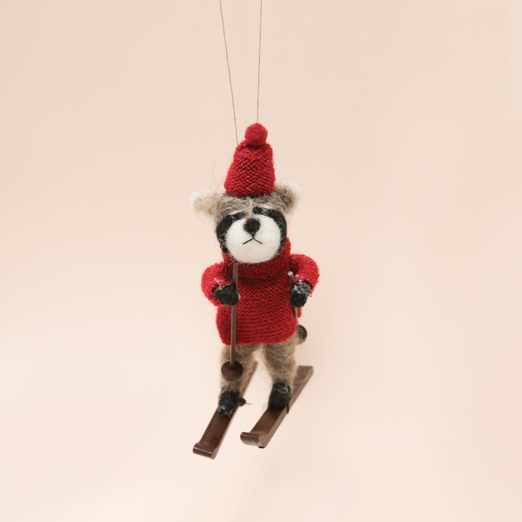 A felt raccoon ornament in a red sweater and hat on brown skis and holding ski poles.