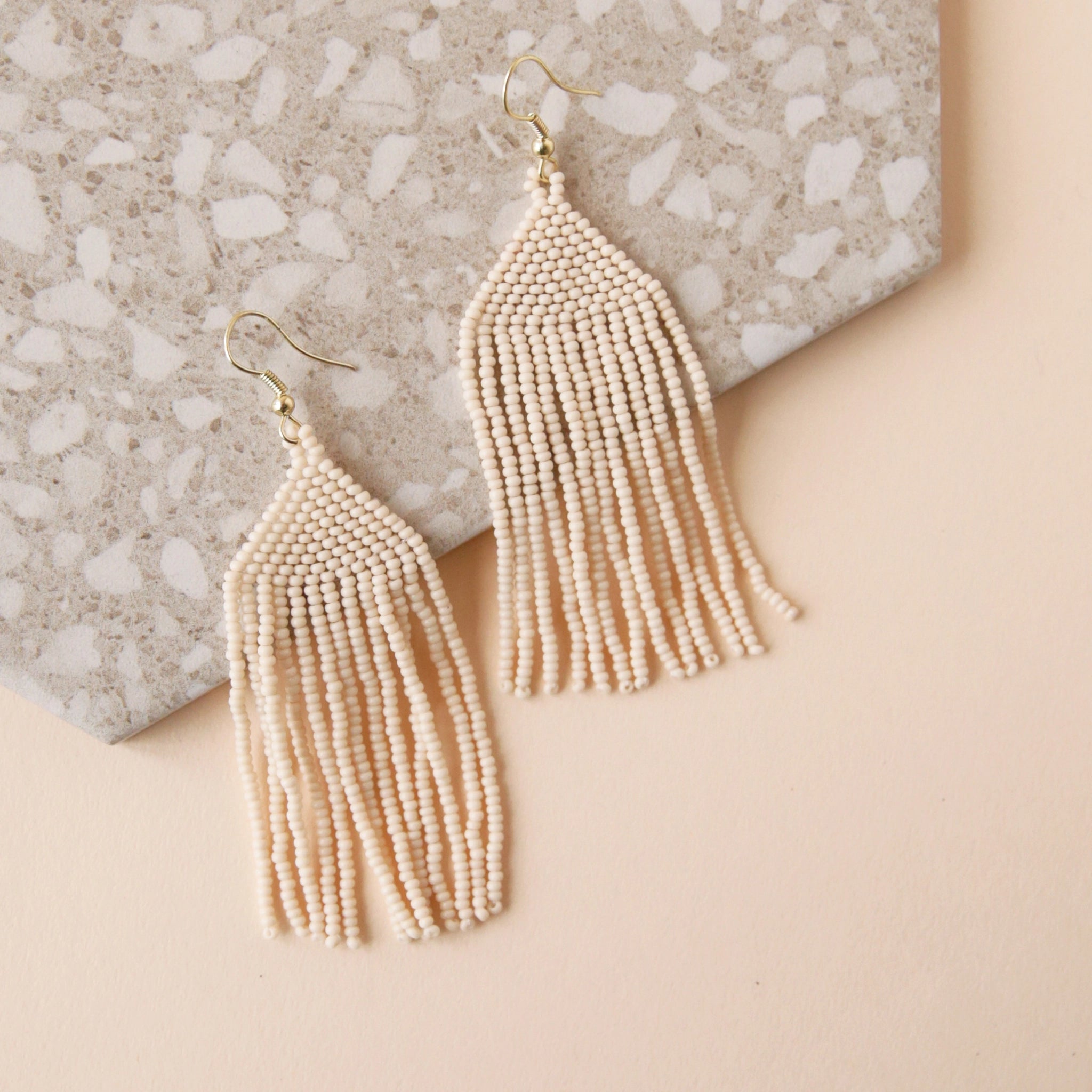 A gold plated hook earring attached to a triangle shaped glass beaded earring in a cream shade along with a fringe detail on the bottom edge.