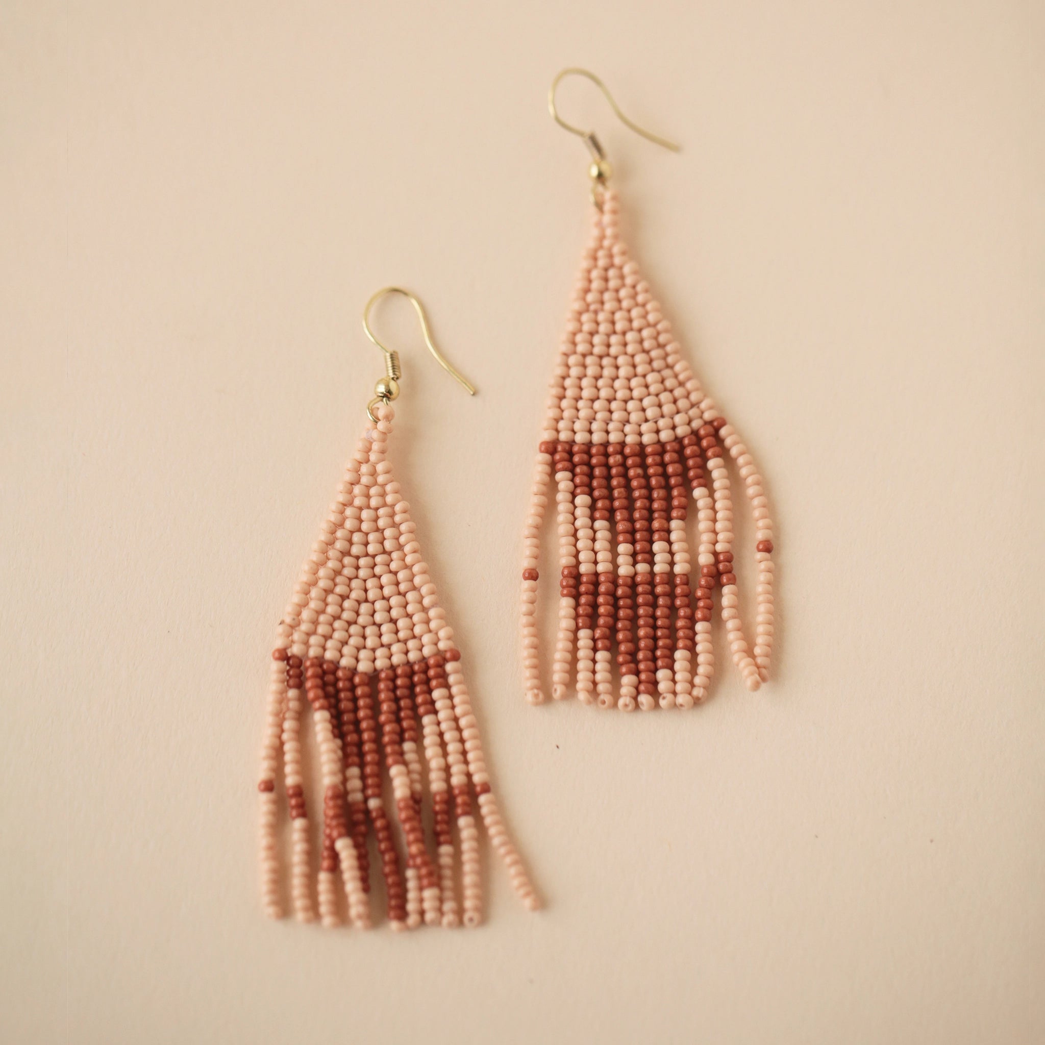 Two triangle shaped beaded earrings with tonal blush and rust and  a modern geometric pattern attached to a gold plated hook style earring.