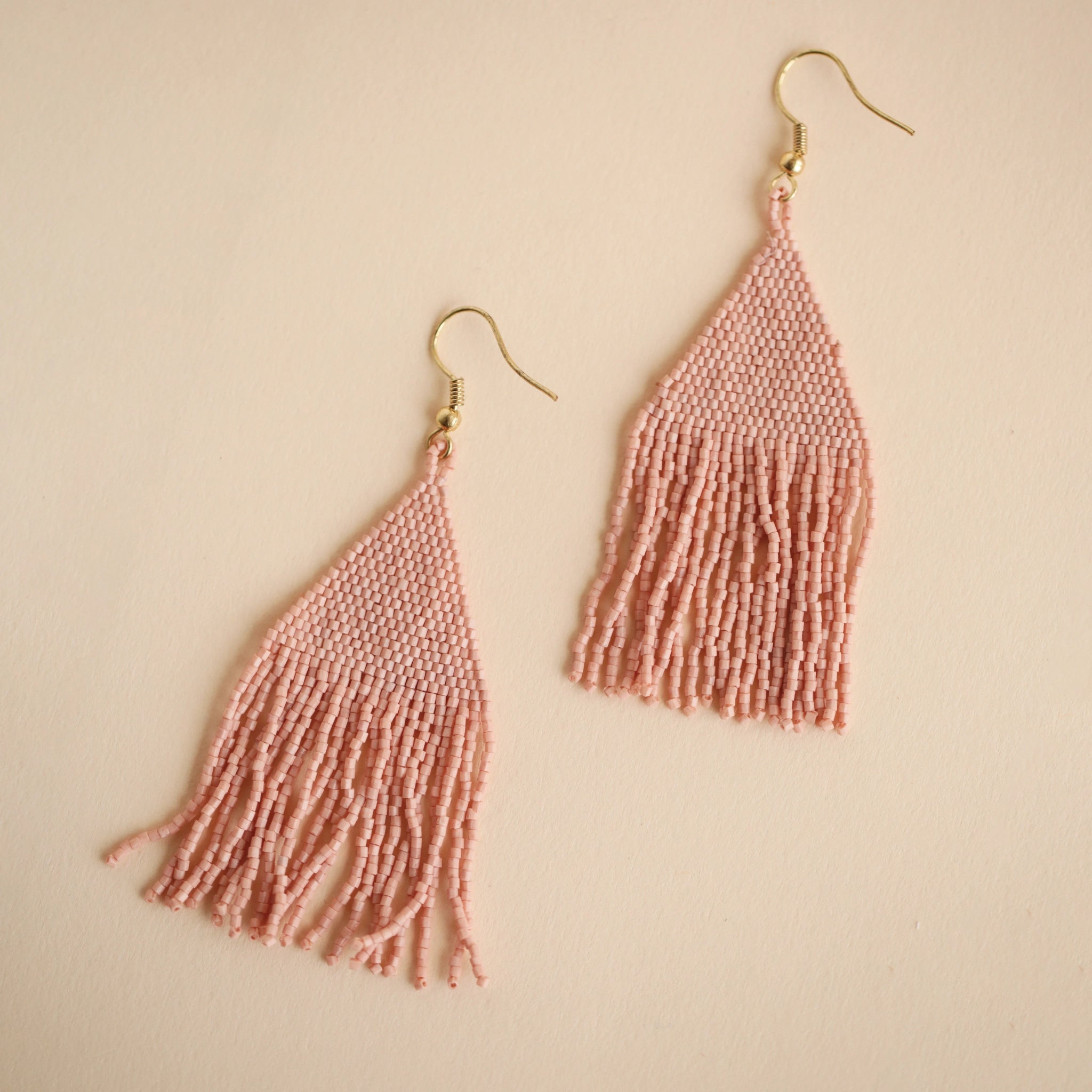 Two blush pink glass beaded earrings with a triangle shape along with fringe detailing on the bottom.