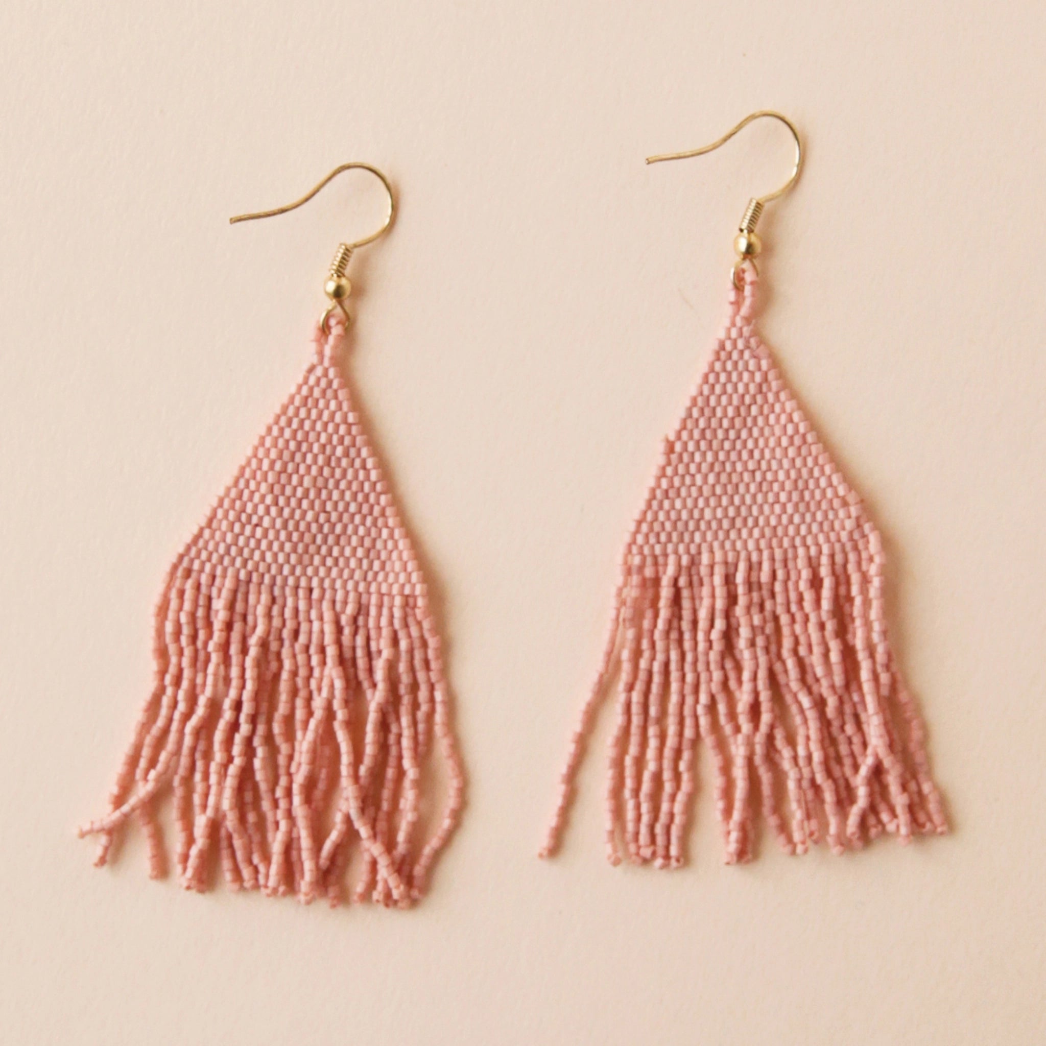 Two blush pink glass beaded earrings with a triangle shape along with fringe detailing on the bottom.