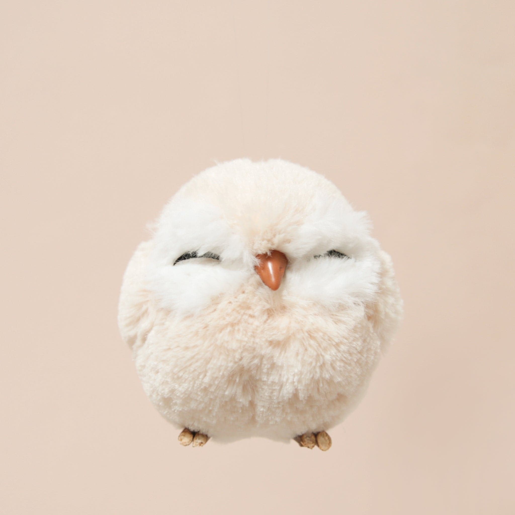 A tan faux fur ball shaped own ornament with two closed eyes, tiny wooden feet and an orange beak.