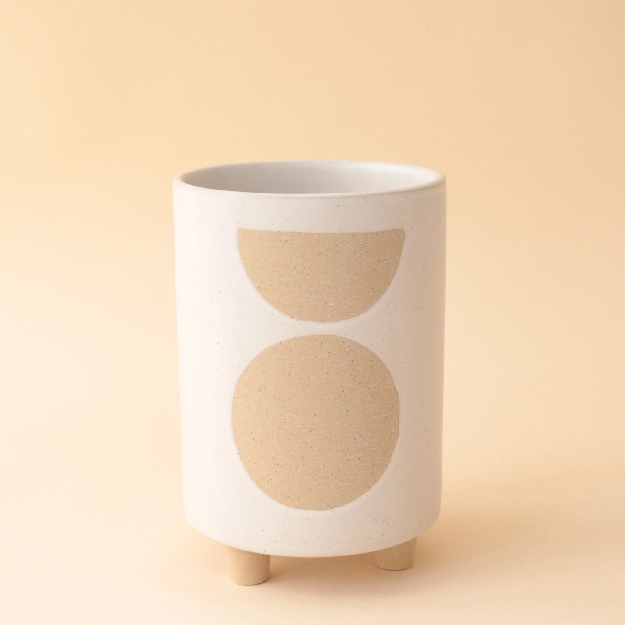 A white ceramic cylinder planter with three small feet on the bottom that elevates it slightly along with a tan circle and half circle design on the front.