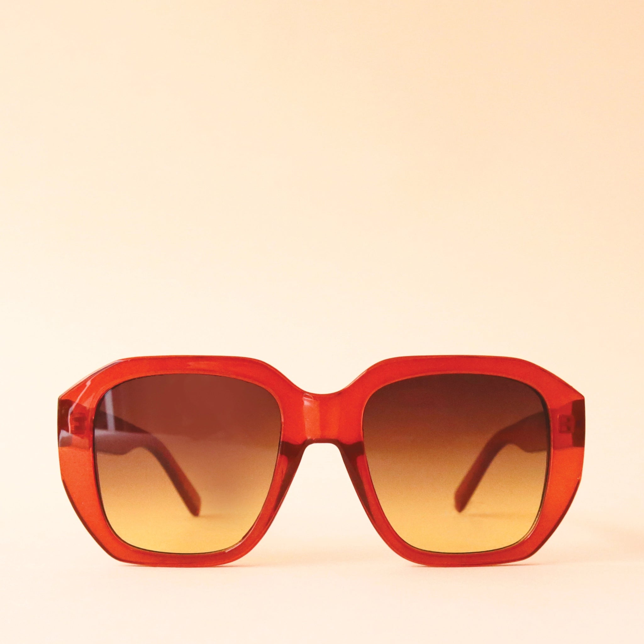 Chunky rust colored sunglasses with a rounded rectangular shape and a brownish gradient lens, photographed in front of a peachy background.