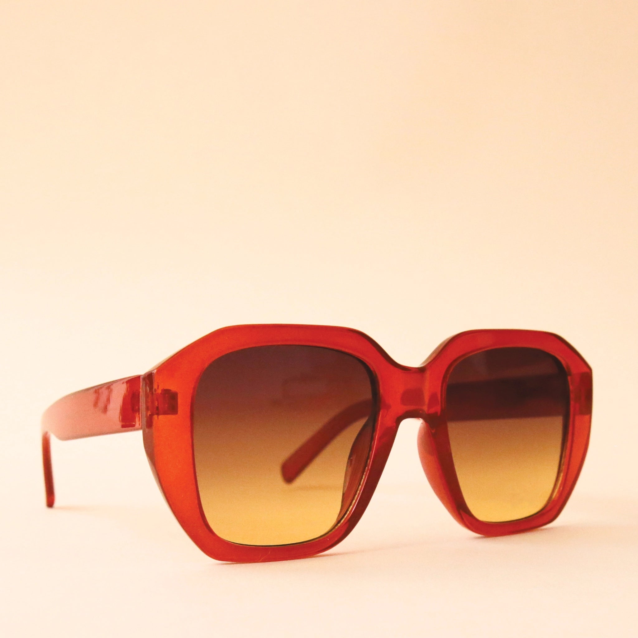 Chunky rust colored sunglasses with a rounded rectangular shape and a brownish gradient lens, photographed in front of a peachy background.