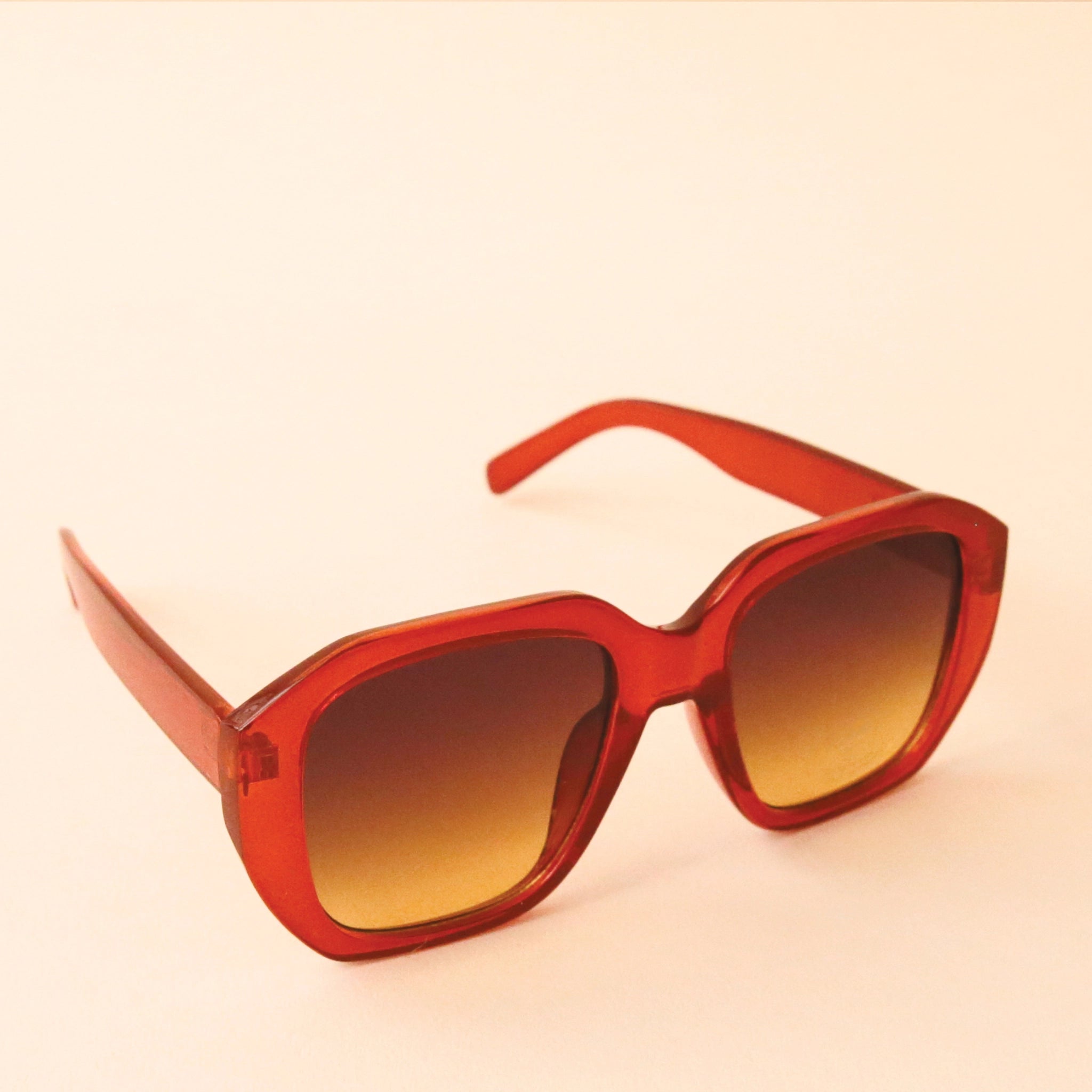 Chunky rust colored sunglasses with a rounded rectangular shape and a brownish gradient lens, photographed in front of a peachy background.