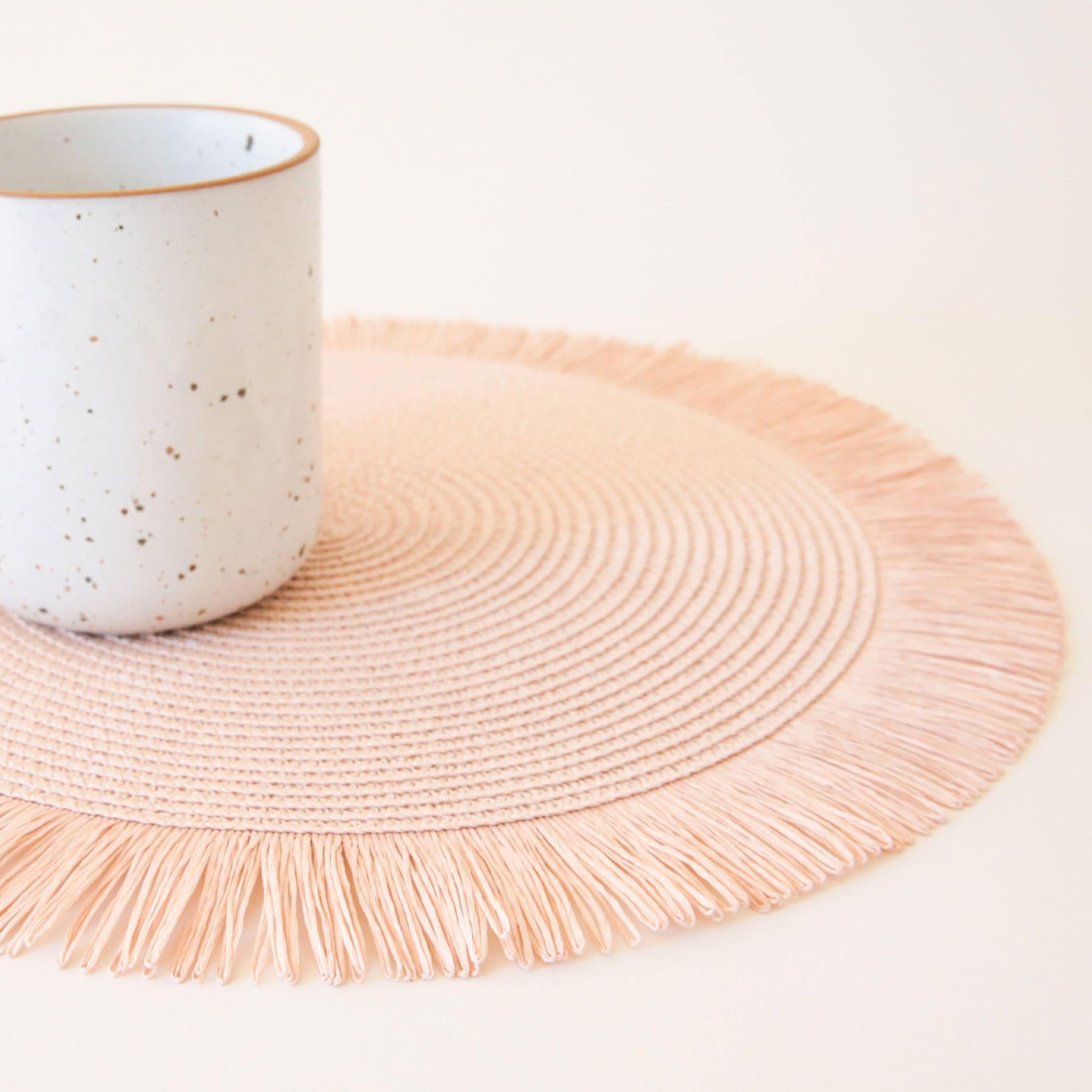 Textured, blush pink circle place mat with bordered in a soft pink fringe. A speckled white mug sits offset on the place mat. 